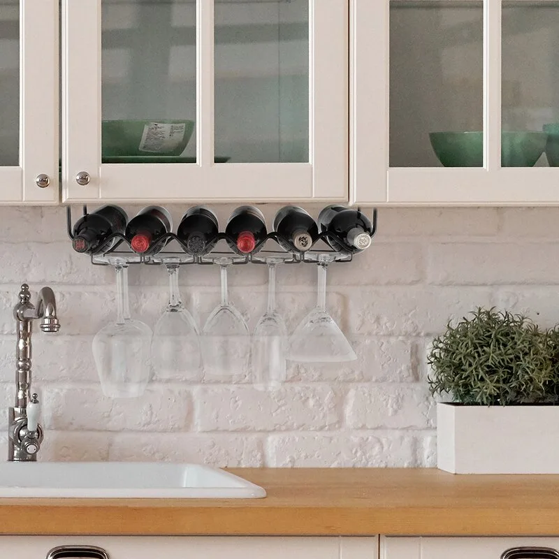 A black metal wine rack holding bottles and hanging glasses is mounted under white kitchen cabinets against a white brick backsplash.