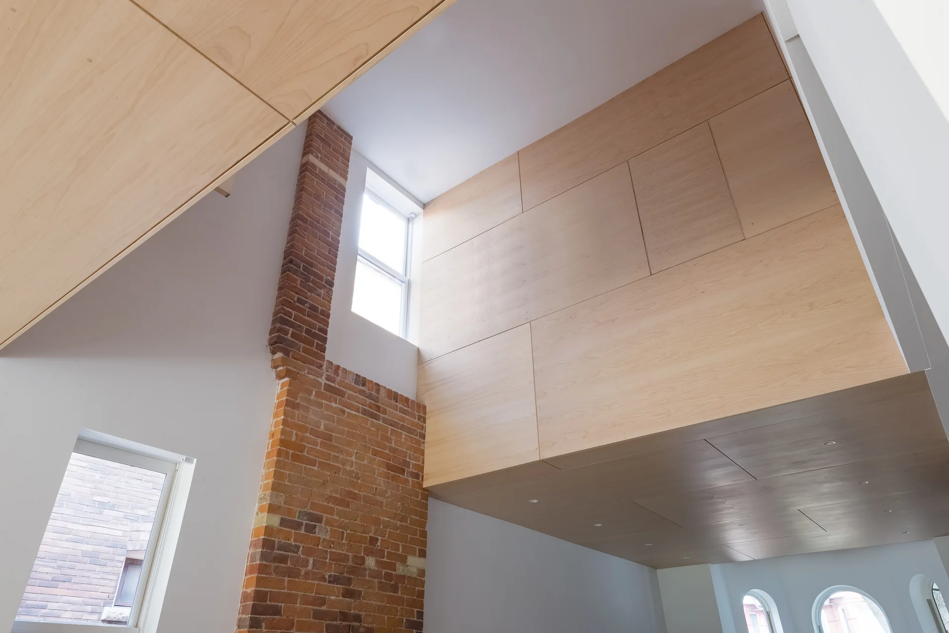 An architectural view of a high-ceilinged loft featuring a red brick column alongside large panels of light wood and white walls with windows.
