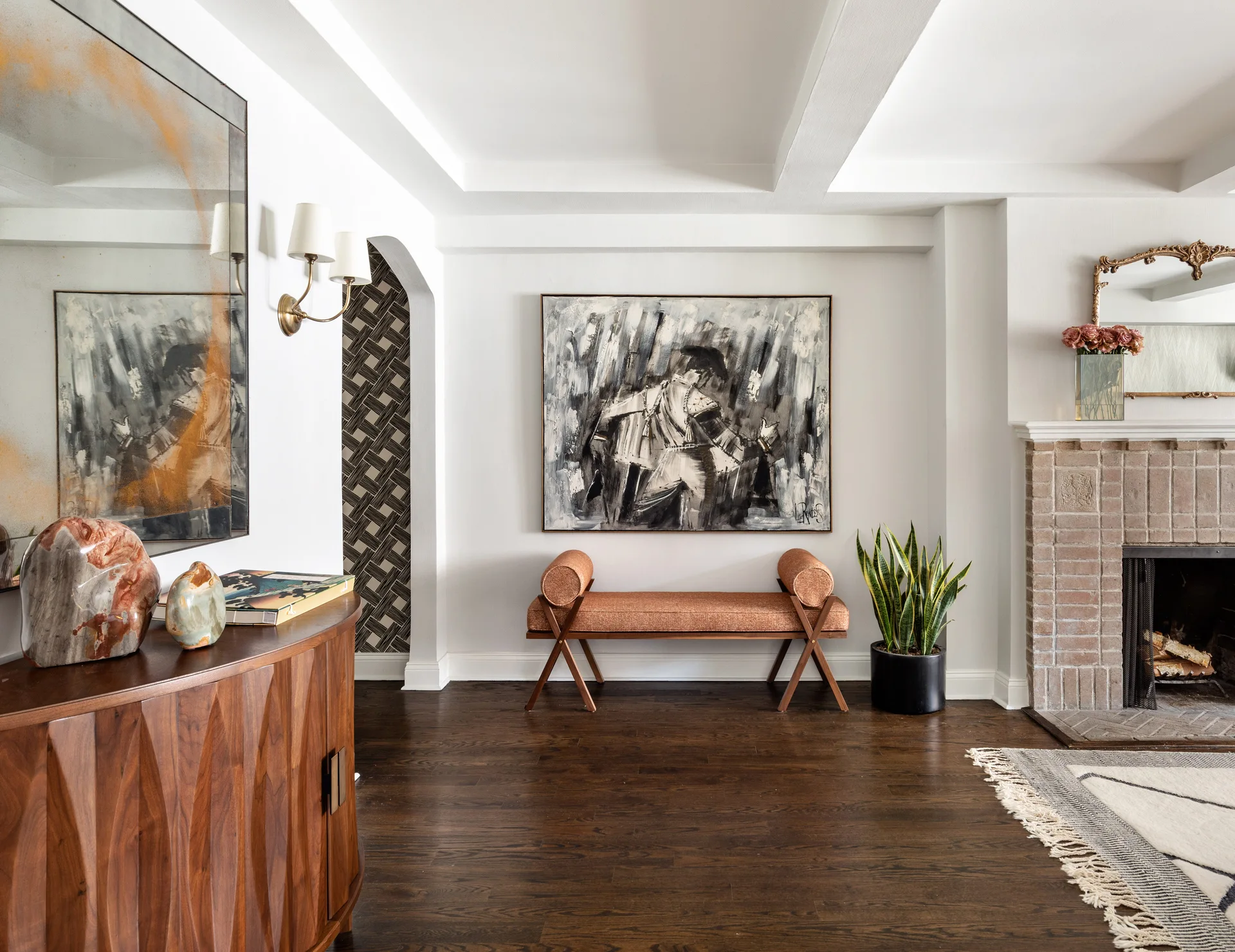 A white living room features a dark wood console table, a terracotta bolster bench under abstract art, and a brick fireplace.