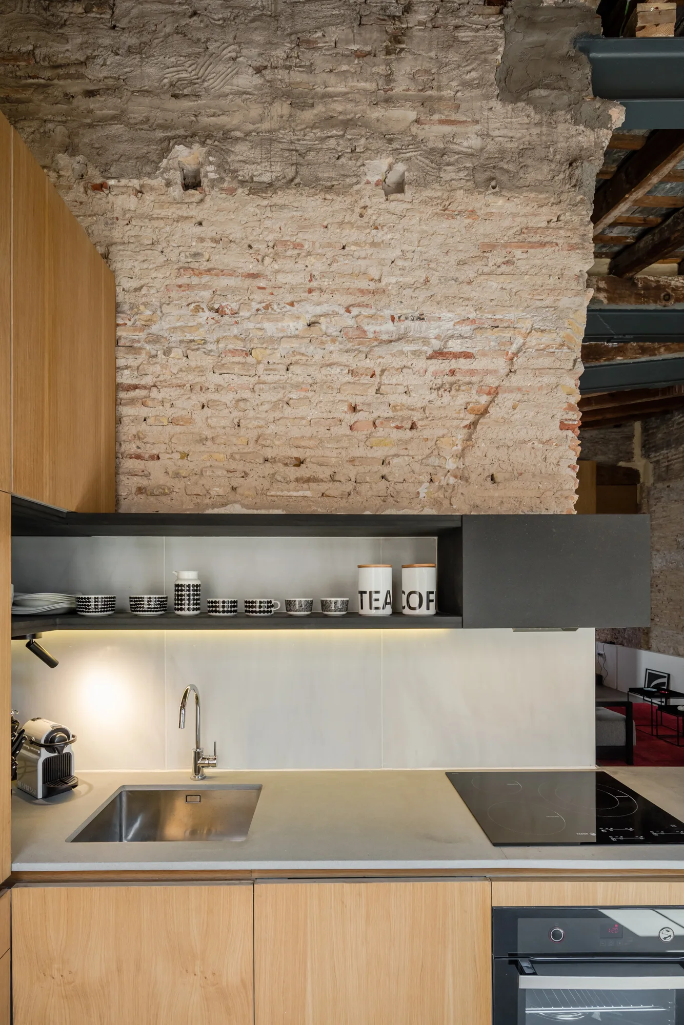 A modern kitchen with light wood cabinetry, a grey stone countertop, and a sleek black floating shelf beneath a raw exposed brick wall.