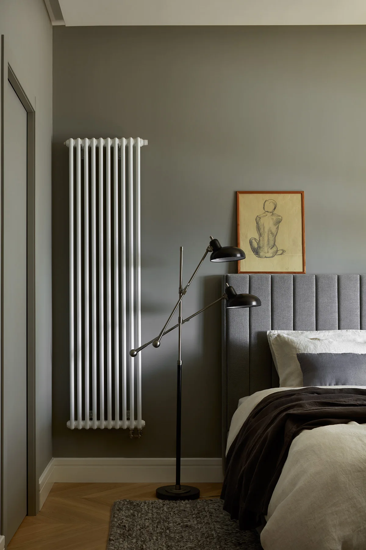 A bedroom corner with sage green walls, a tall white column radiator, a black floor lamp, and a grey upholstered headboard.
