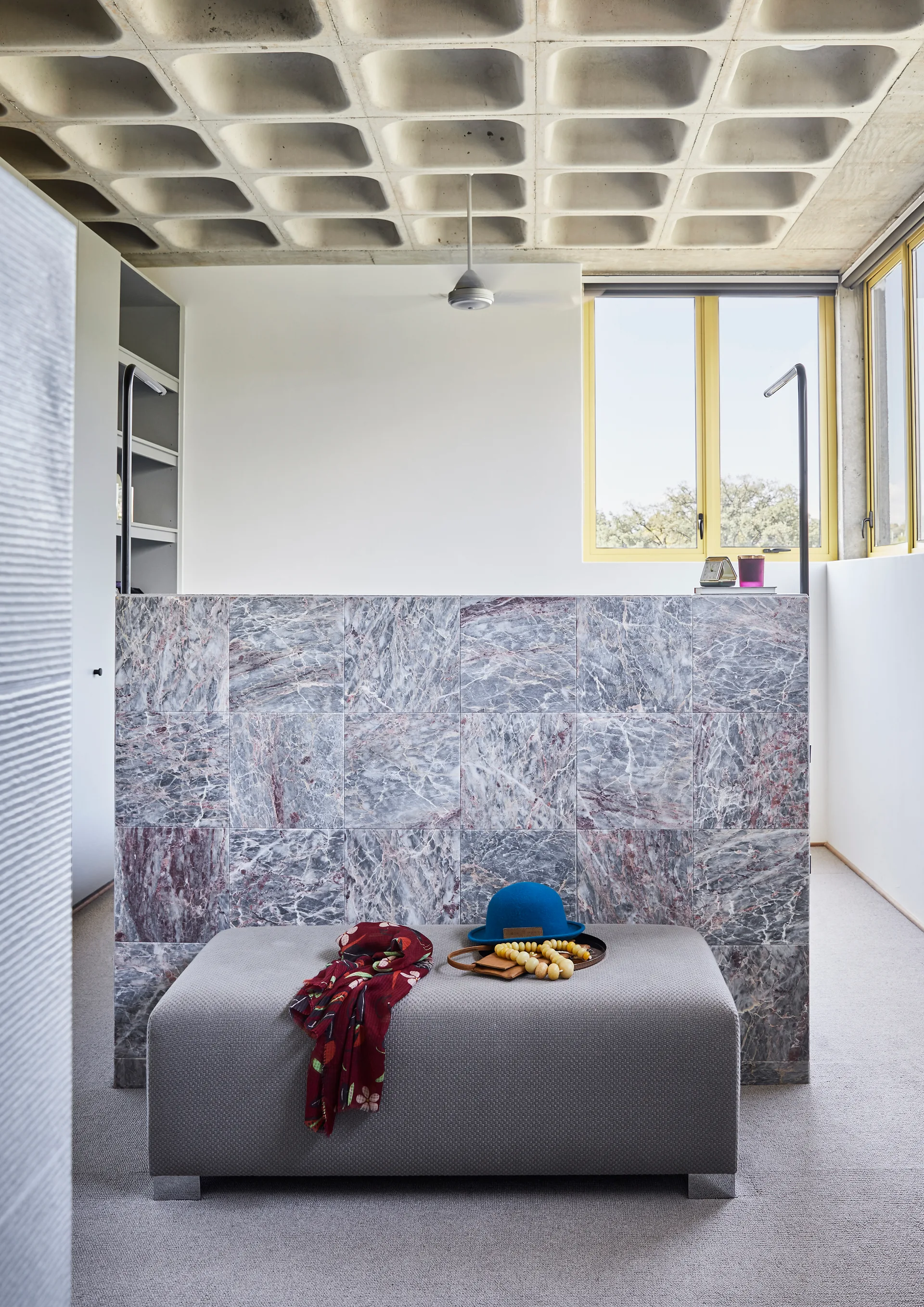 An upholstered gray bench sits in front of a purple-veined marble partition wall under a concrete waffle ceiling with yellow-framed windows.