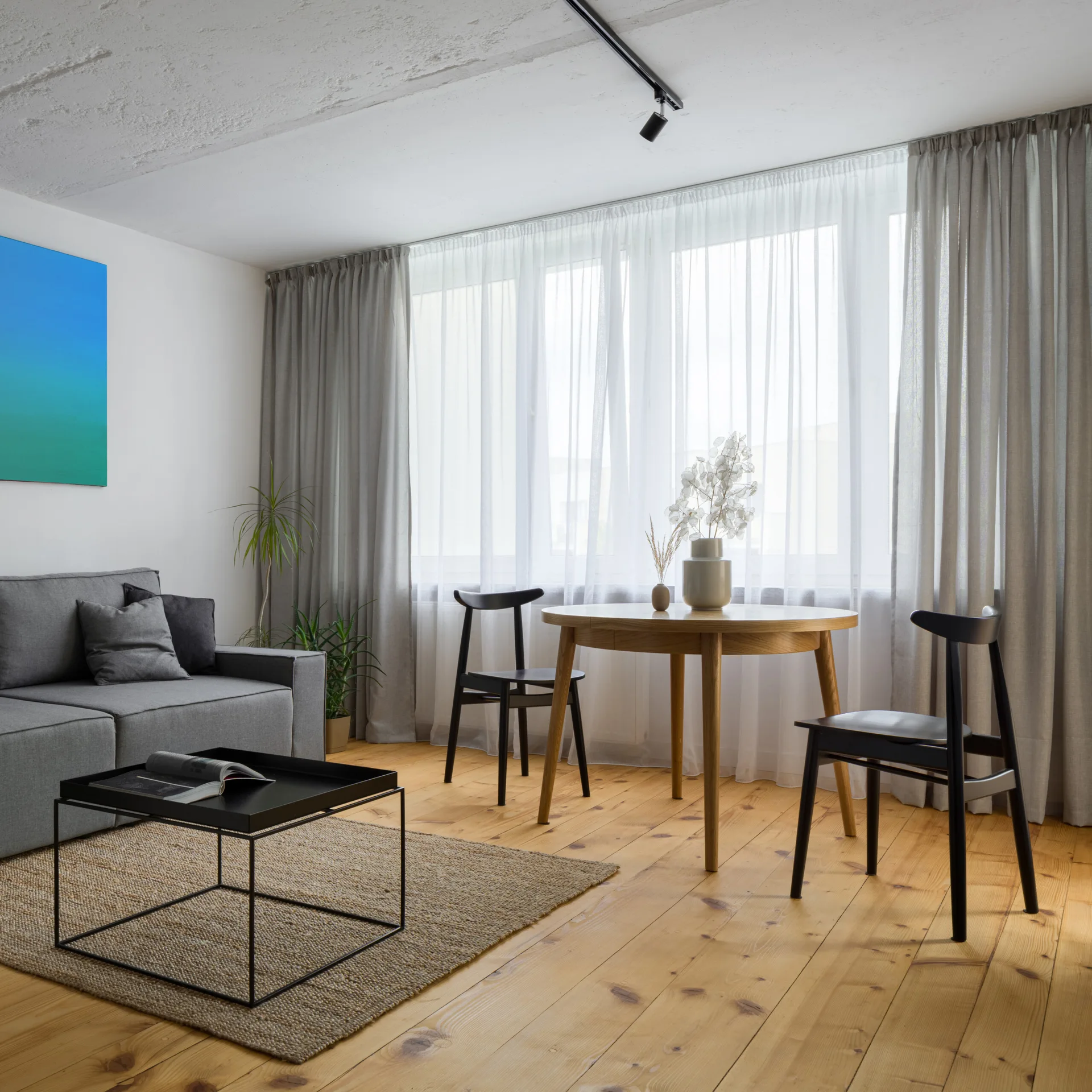 A bright living and dining room features a grey sofa, a minimalist black coffee table on a jute rug, and a round wooden dining table with black chairs.