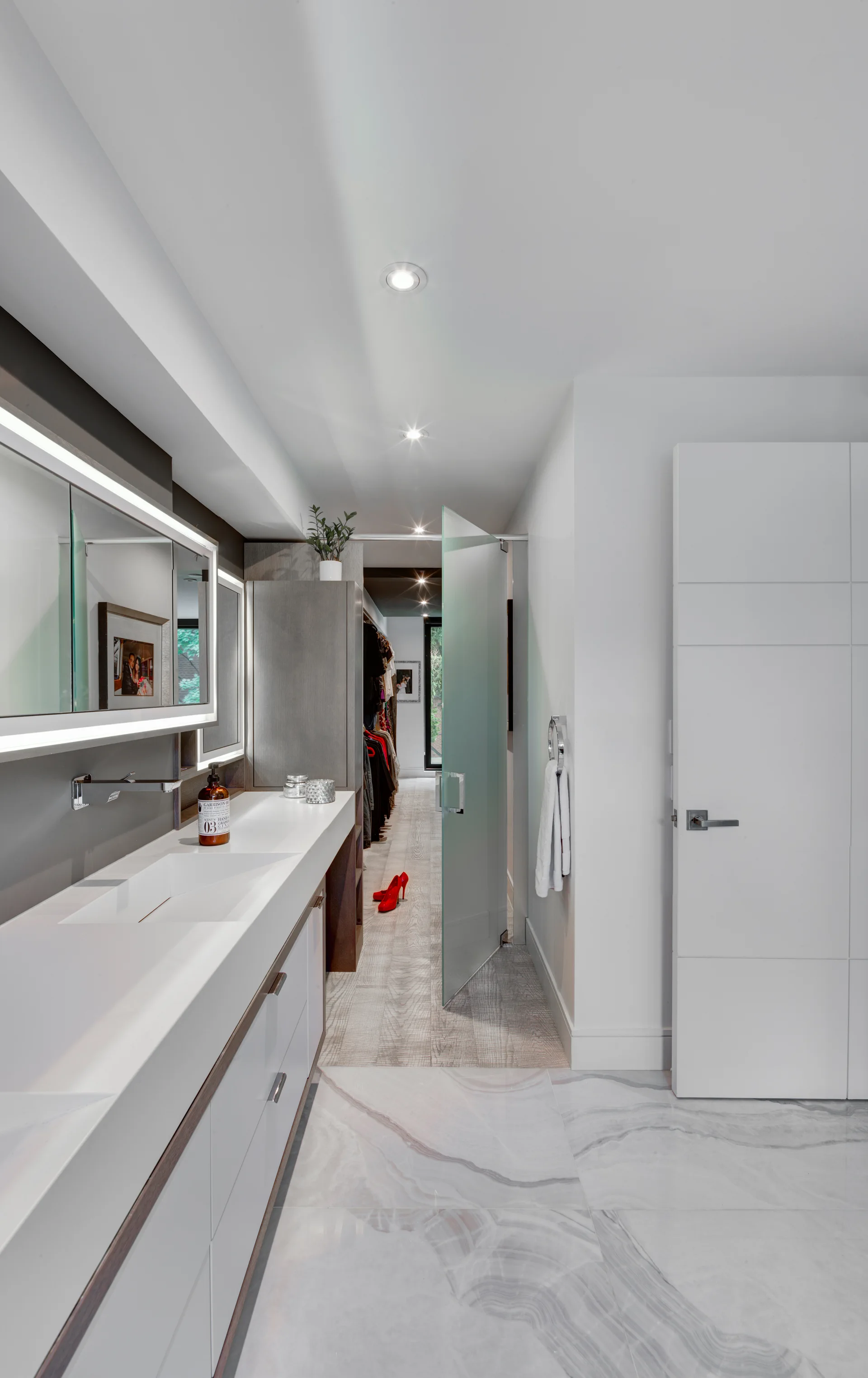 A modern bathroom features a long white vanity with an integrated sink, a backlit mirror, and a frosted glass door leading to a walk-in closet.