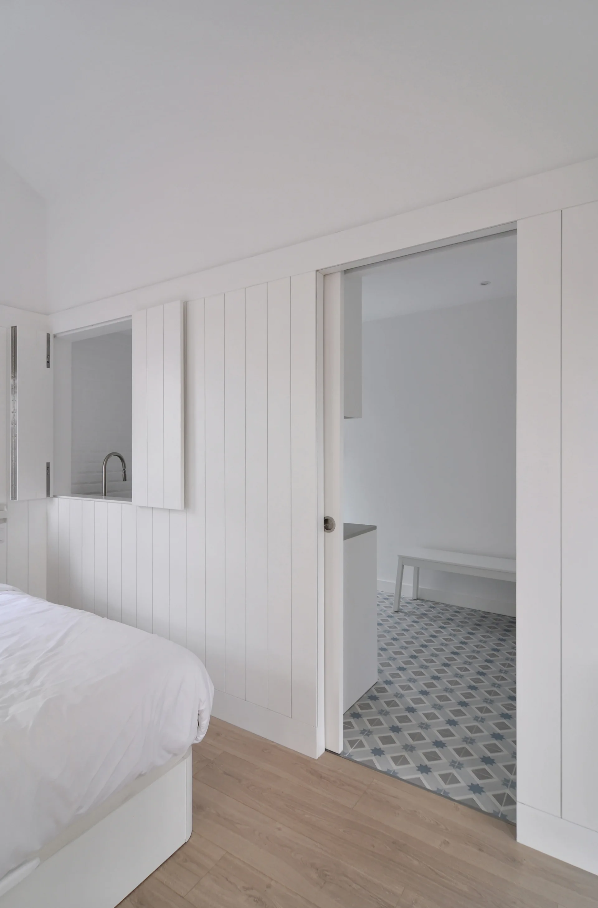 A minimalist white bedroom featuring vertical wood wall paneling, a sliding pocket door, and a small shuttered interior window.