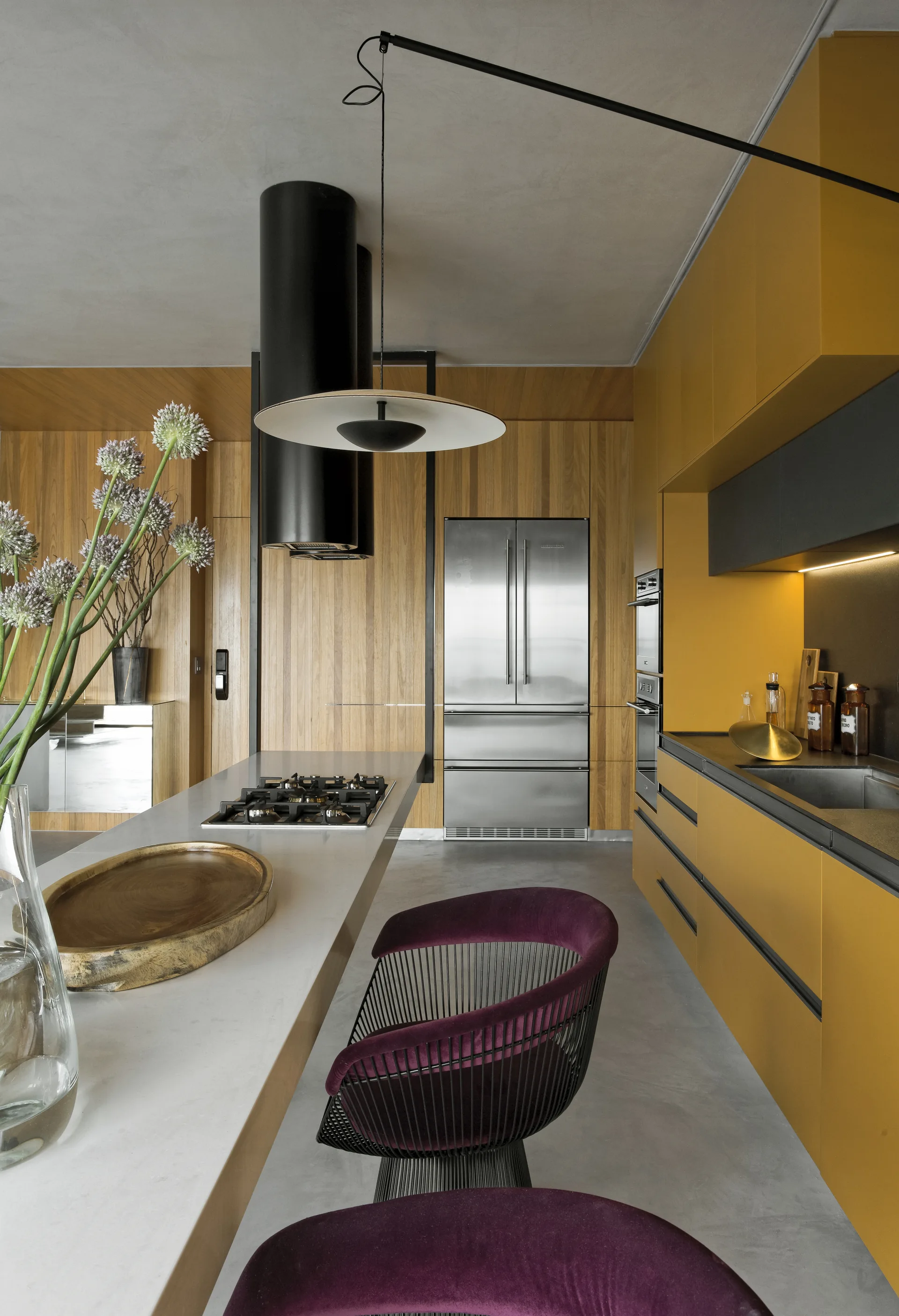 Modern kitchen with mustard yellow cabinets, wood slatted walls, a grey island with purple bar stools, and stainless steel appliances.