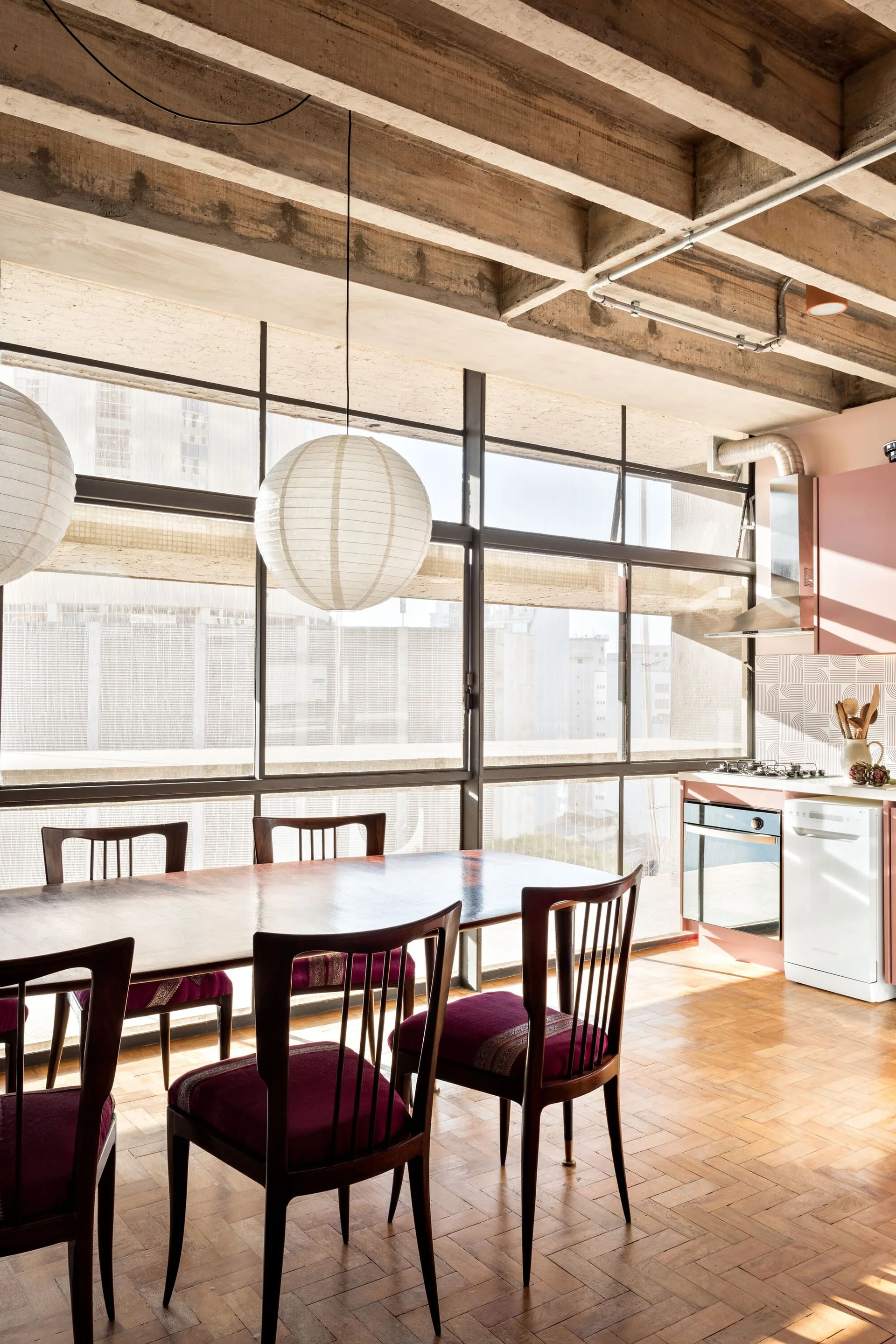 A dining room features a wooden table and chairs beneath paper globe pendant lights and an exposed concrete beamed ceiling next to pink kitchen cabinets.