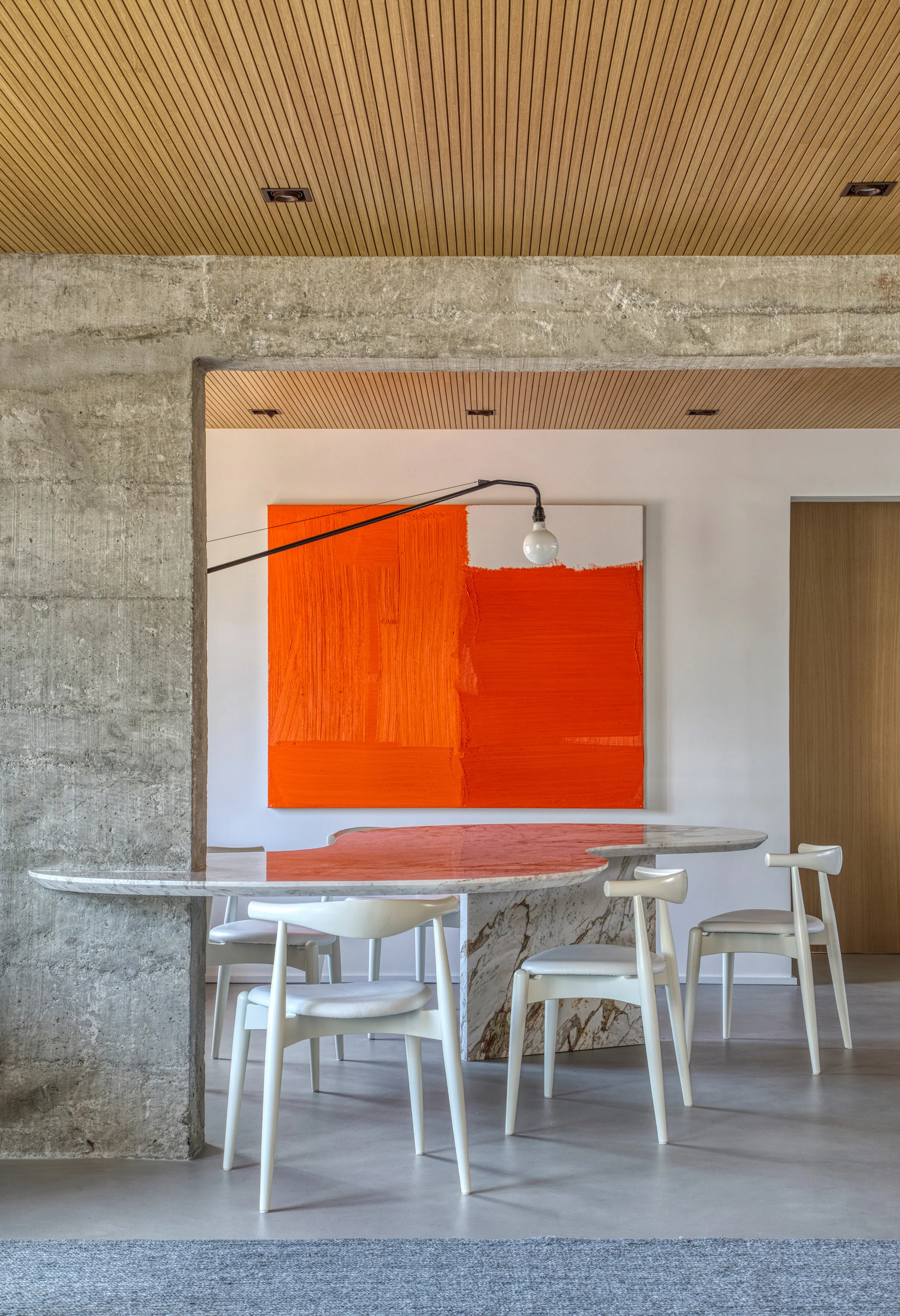 A modern dining area features a sculptural marble table with white wooden chairs beneath a slatted wood ceiling, set next to a raw concrete pillar.