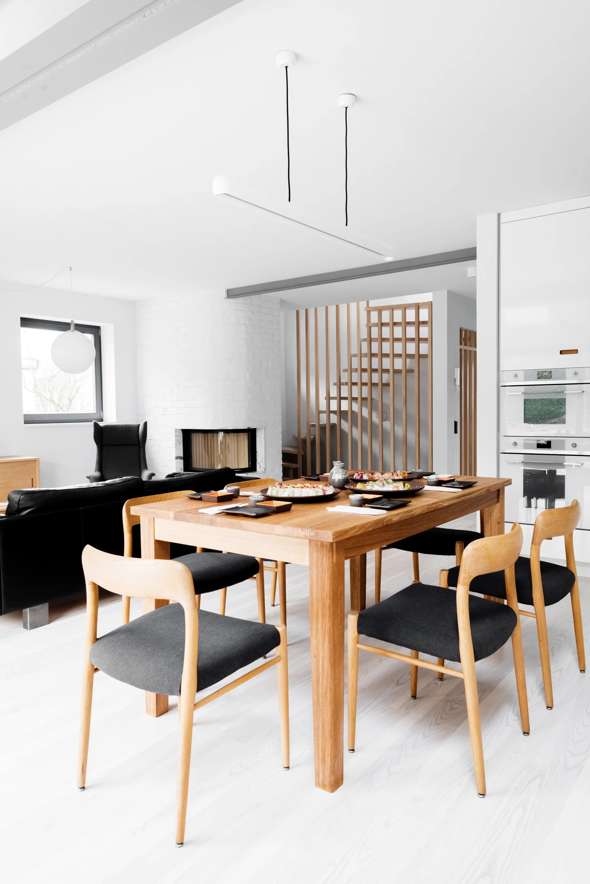 A bright dining room featuring a wooden table and chairs with dark cushions, set against a white fireplace and a slatted wood staircase.