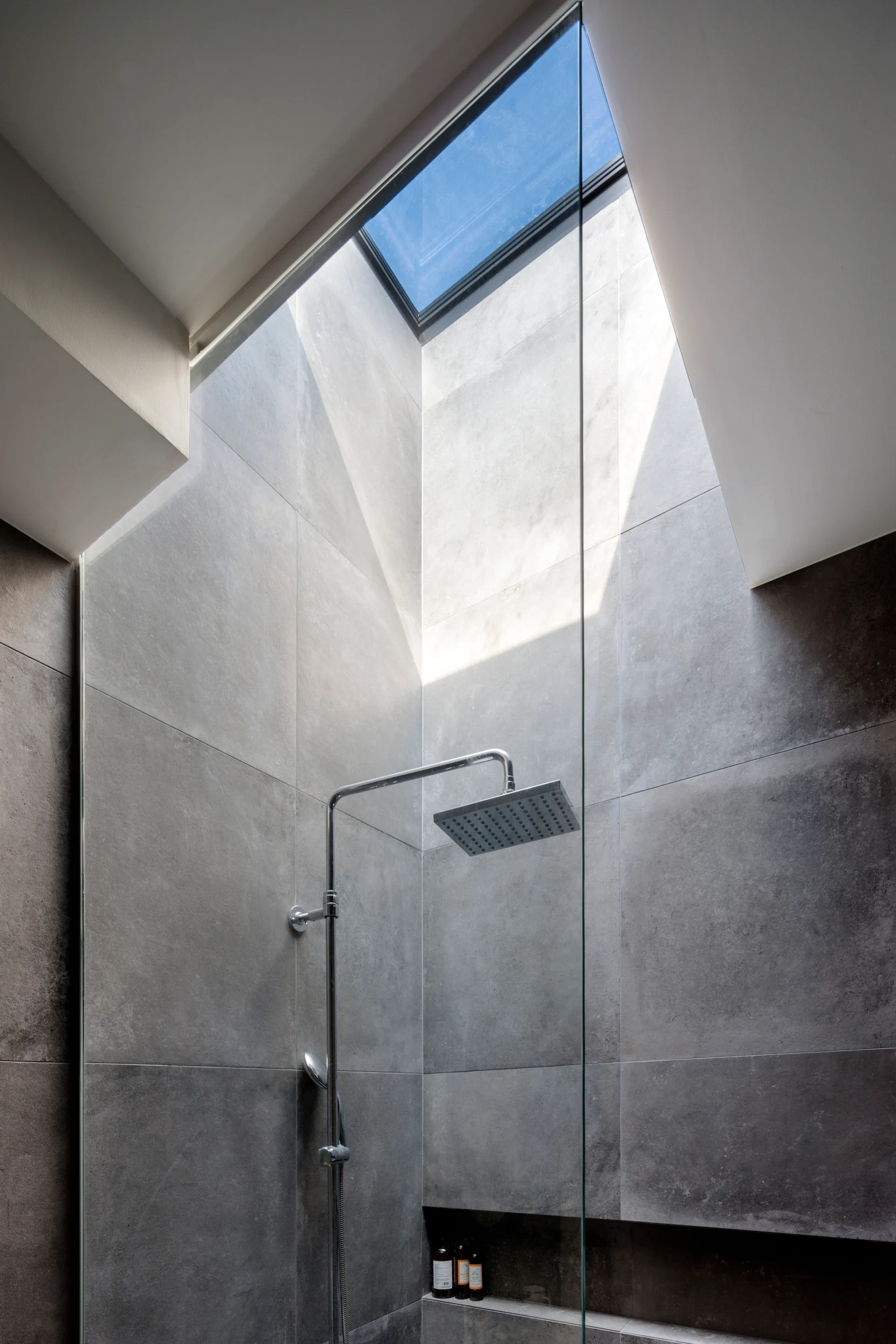A modern walk-in shower features large-format grey wall tiles, a square chrome shower head, a glass partition, and a rectangular ceiling skylight.