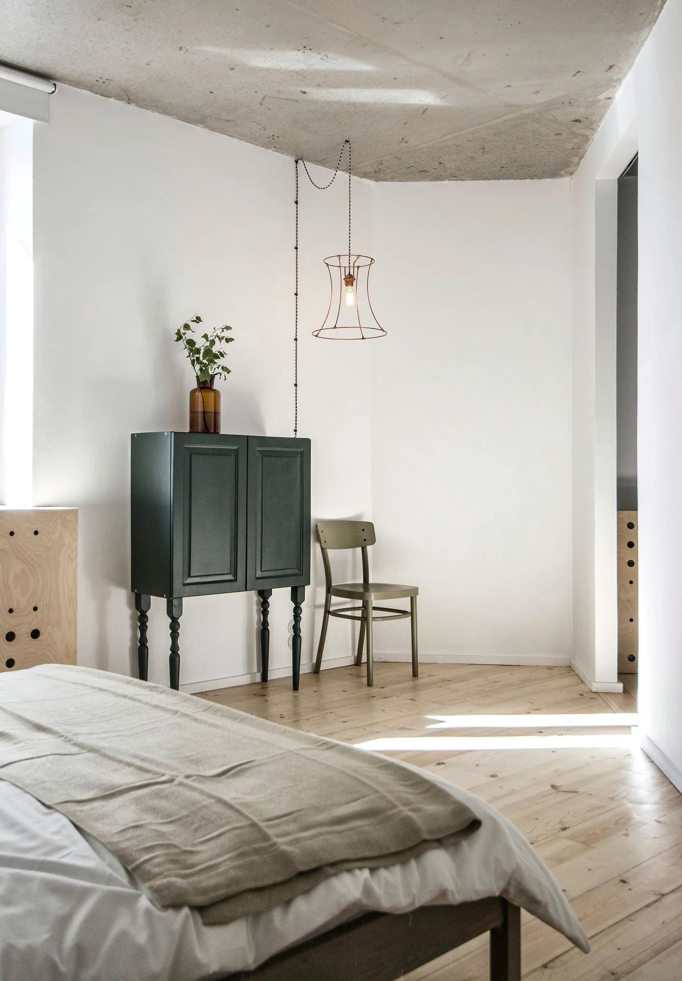 A bedroom corner features a dark green cabinet, a wooden chair, and a wire pendant light beneath a raw concrete ceiling and light wood floors.