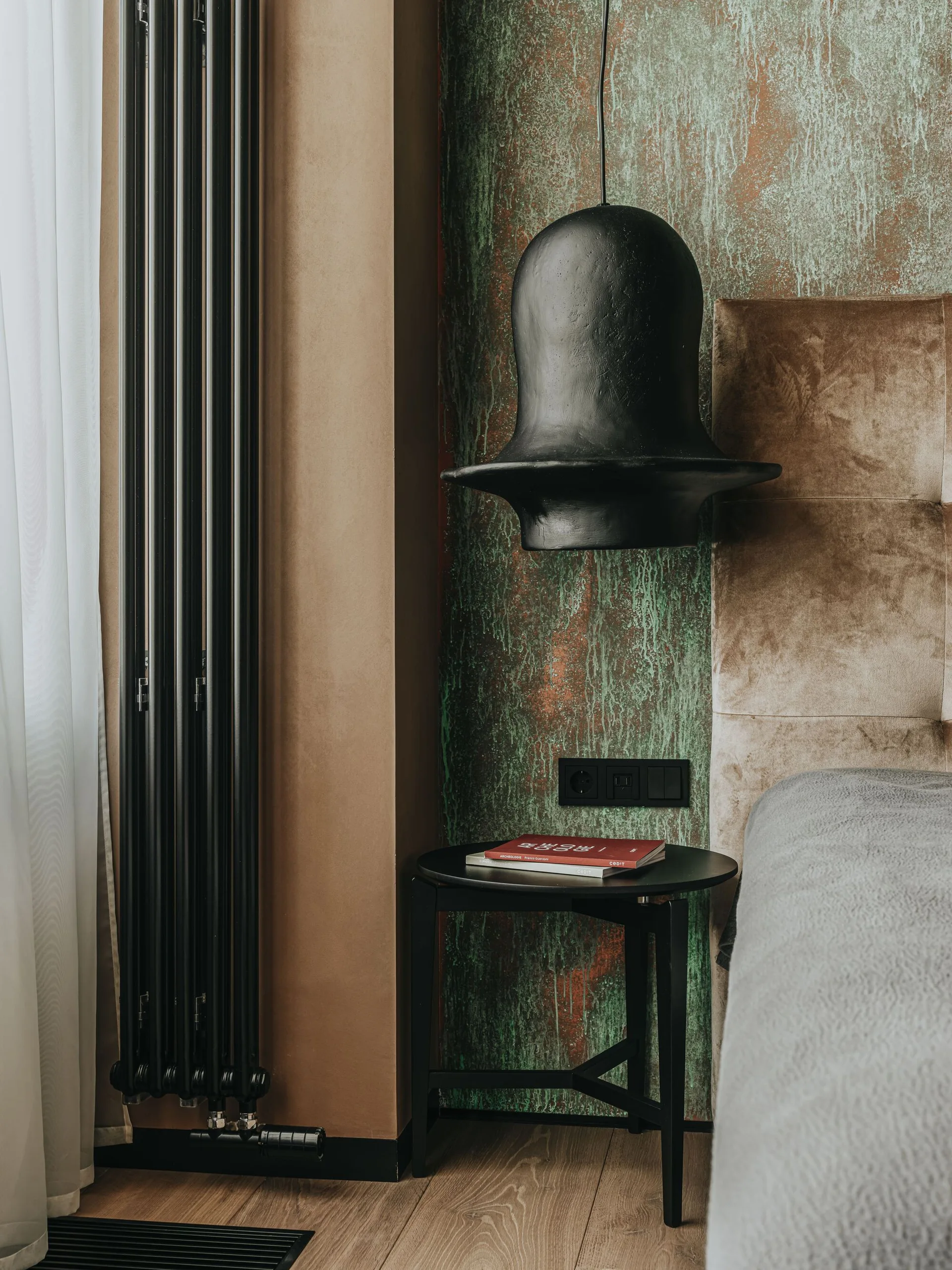 A bedroom bedside corner featuring a textured green oxidized accent wall, a black sculptural pendant light, and a plush upholstered headboard.