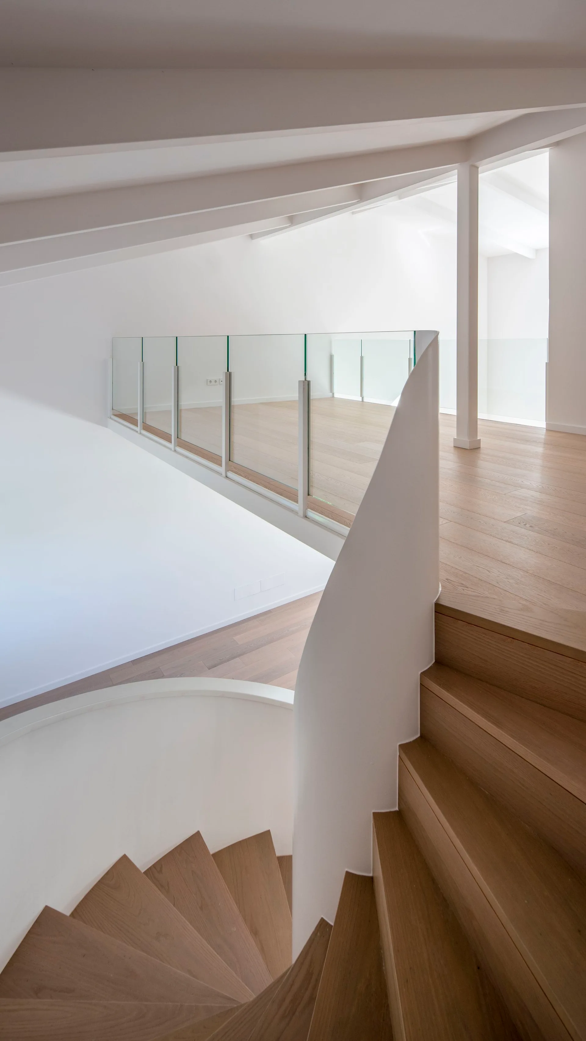 A curved oak staircase leads to a bright white mezzanine featuring light wood flooring and a clear glass panel railing.