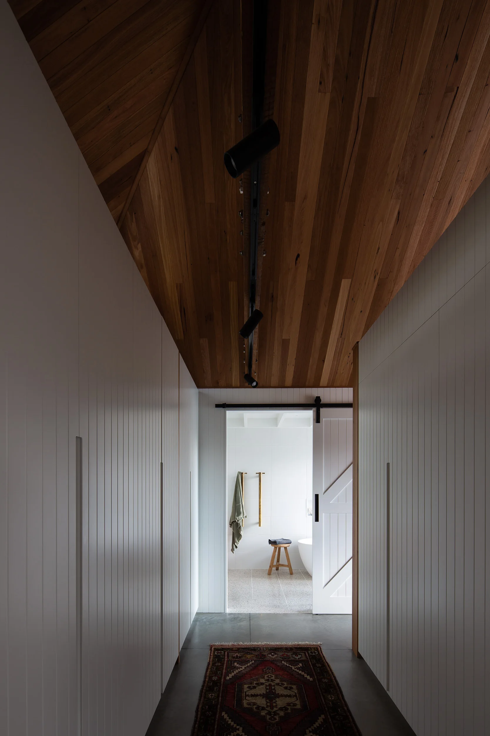 A narrow hallway with white built-in storage cabinets, a sloped wood plank ceiling, and a traditional runner rug leads to a bright bathroom.