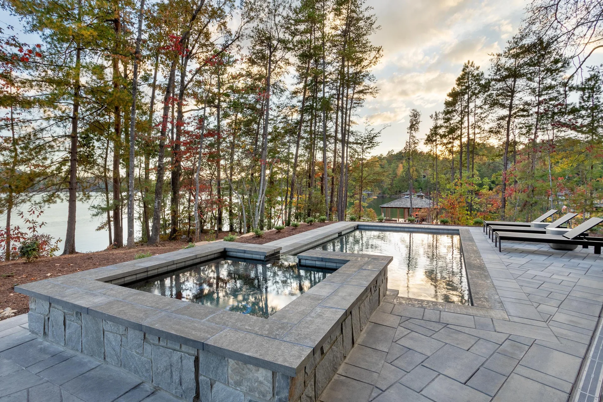 An outdoor swimming pool and raised square spa on a grey stone patio overlooking a lake surrounded by tall pine trees.