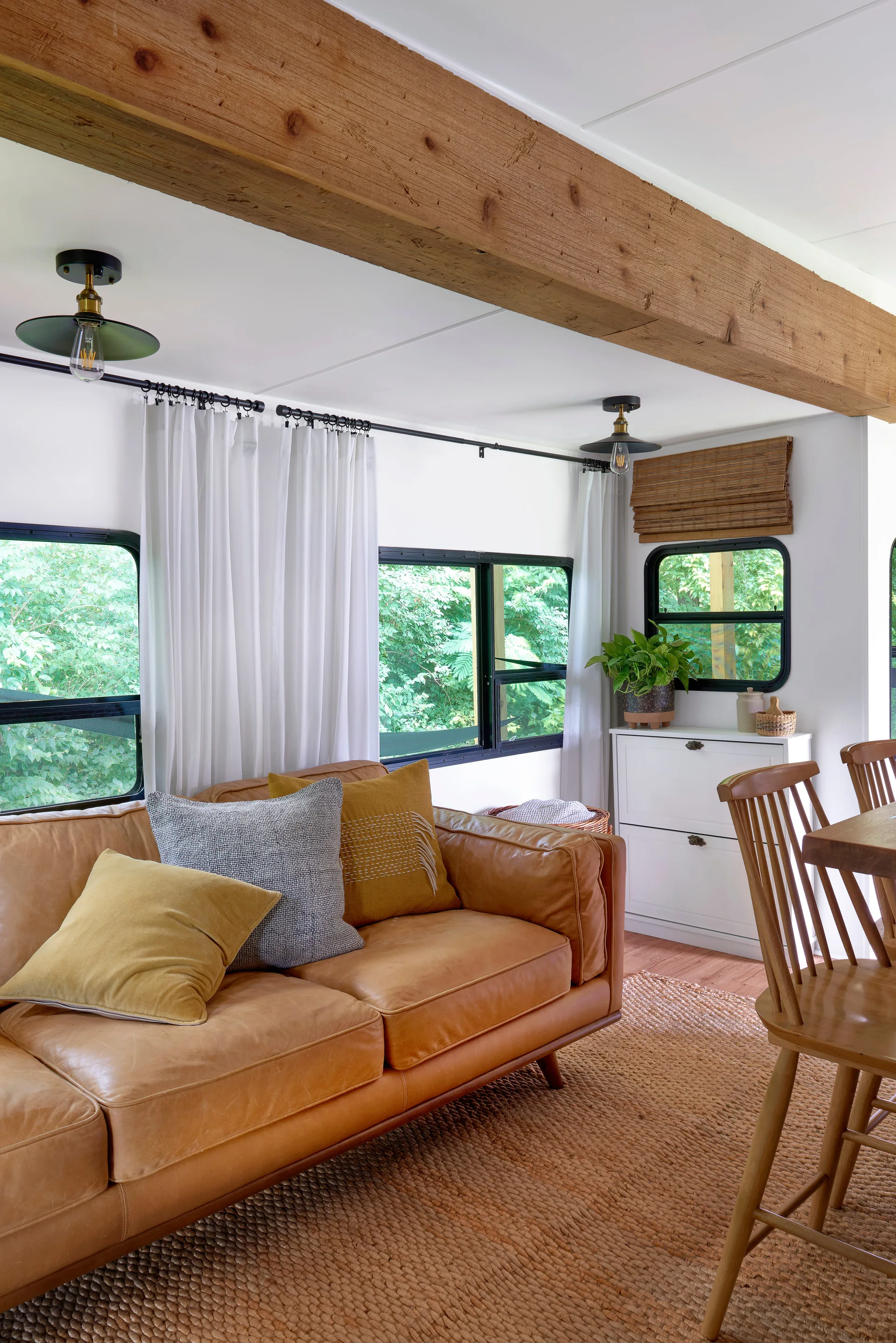 A tan leather sofa with throw pillows sits in a bright room beneath an exposed wood ceiling beam and next to a wooden dining chair.