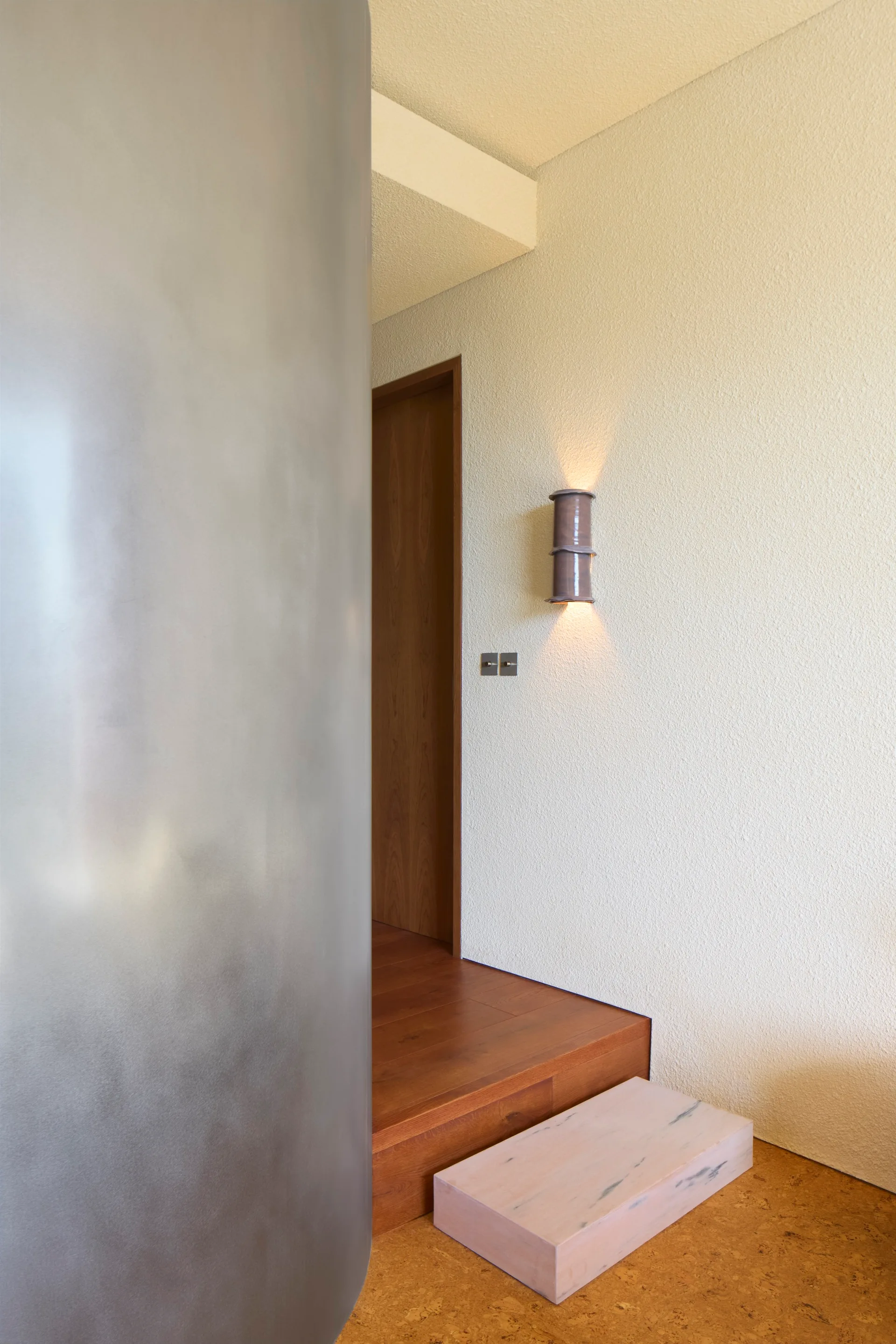 A minimalist hallway transition featuring a curved silver partition, textured cream walls, a stepped wood platform, and a glowing cylindrical wall sconce.
