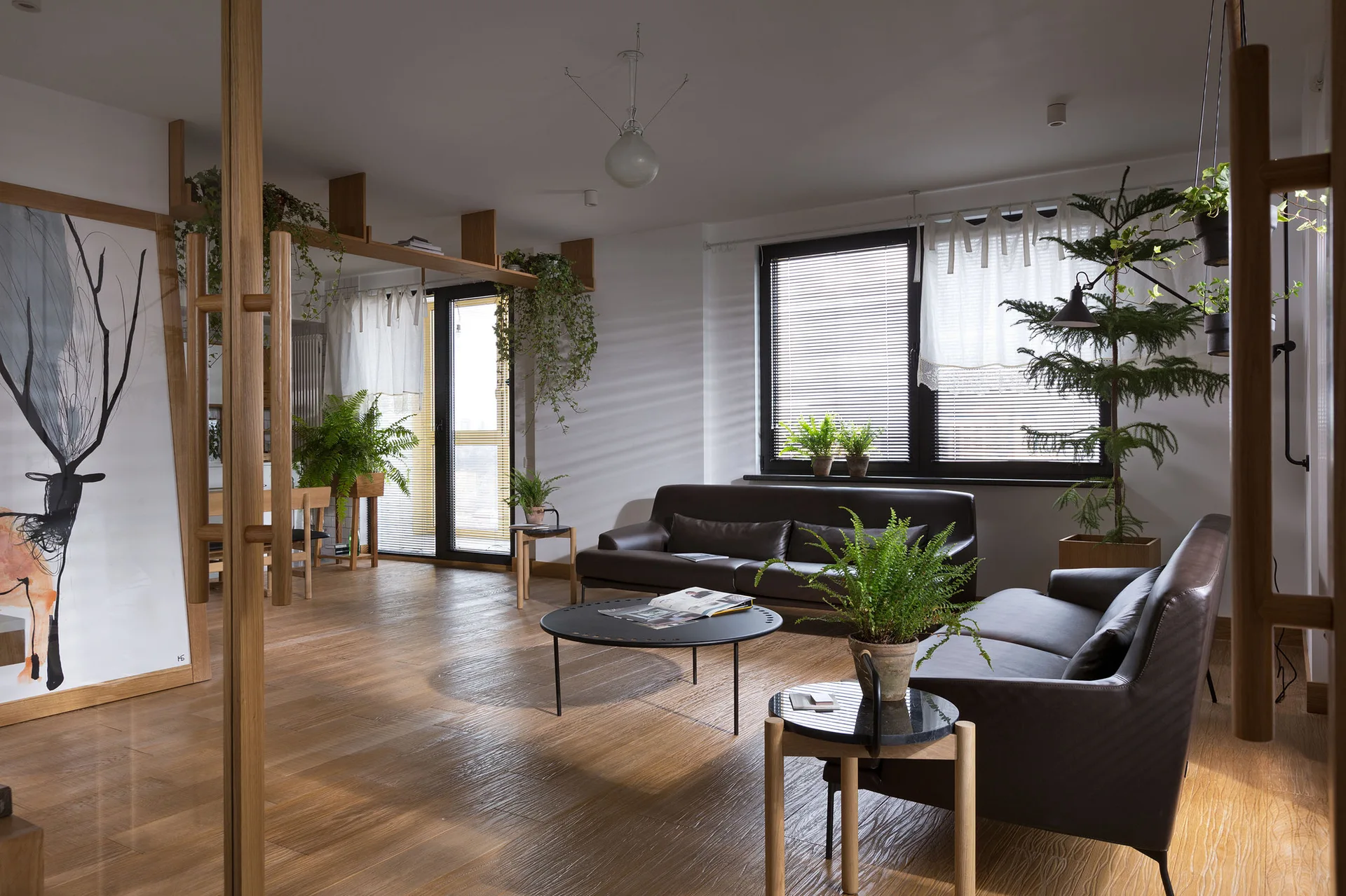 A modern open-plan living room with dark brown leather sofas, wood flooring, a round black coffee table, and multiple potted plants by a large window.