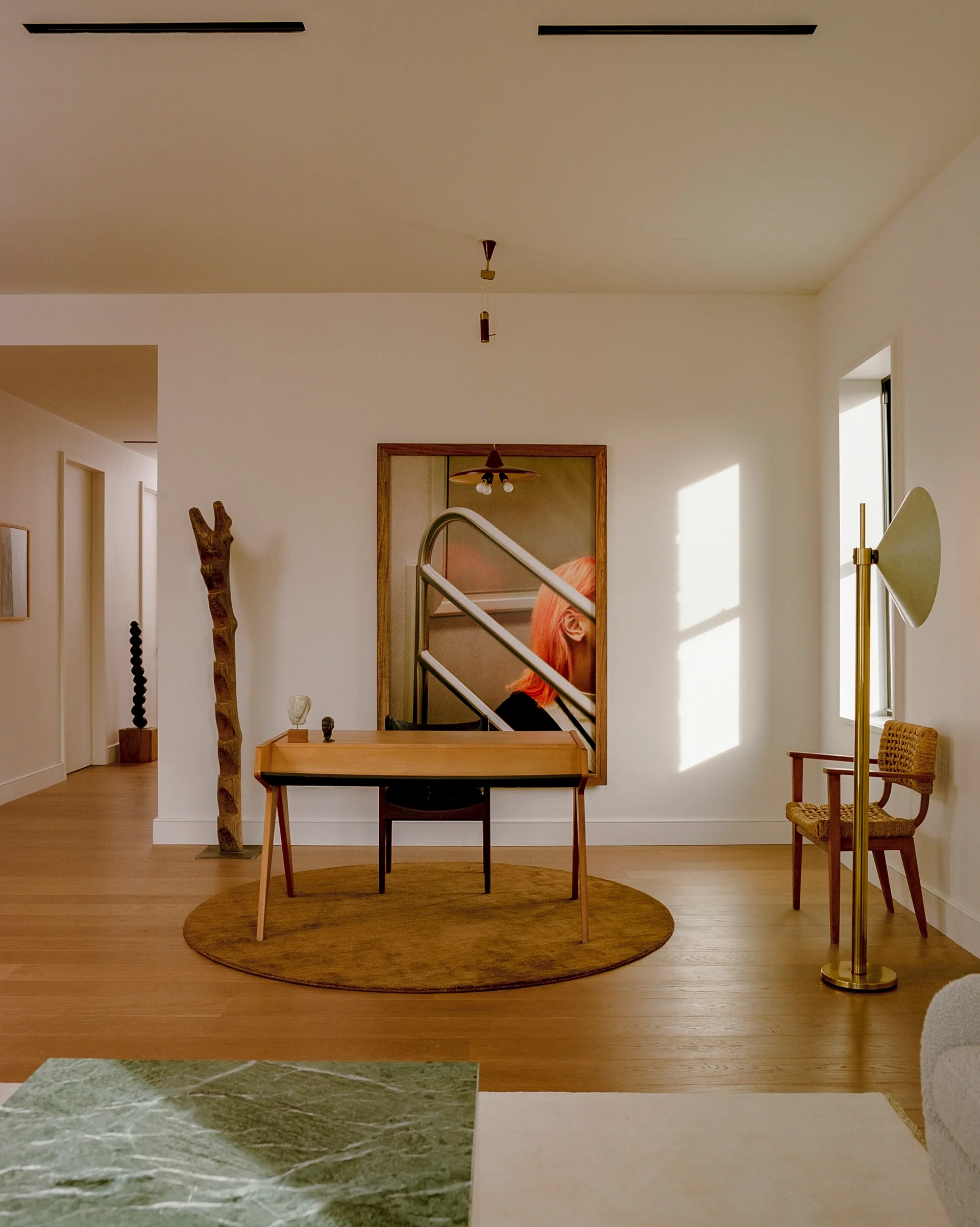 A wooden desk sits on a round brown rug in a minimalist office with white walls, a large framed artwork, and a brass floor lamp.