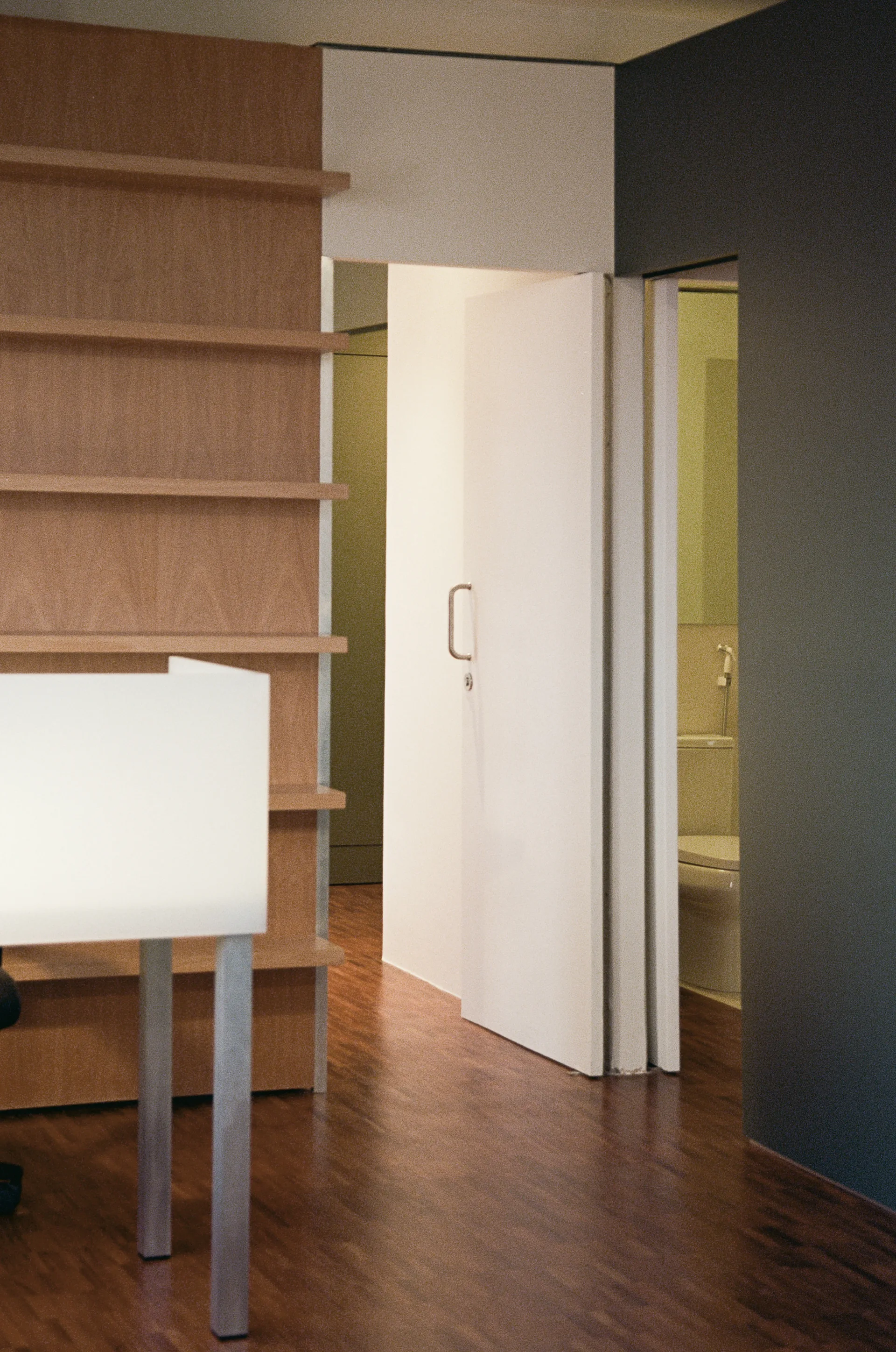 An interior space with a built-in wood shelving unit, a white door opening into a bathroom, and a dark grey accent wall over wooden floors.