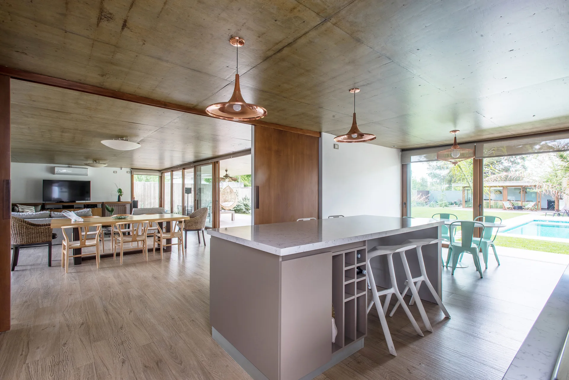 A modern kitchen and dining area features a taupe island with copper pendant lights under an exposed concrete ceiling looking out to an outdoor pool.