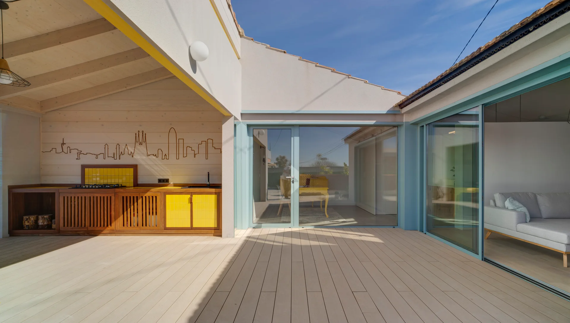 A modern wood-decked patio with a covered outdoor kitchen featuring yellow cabinets and large glass sliding doors framed in teal.