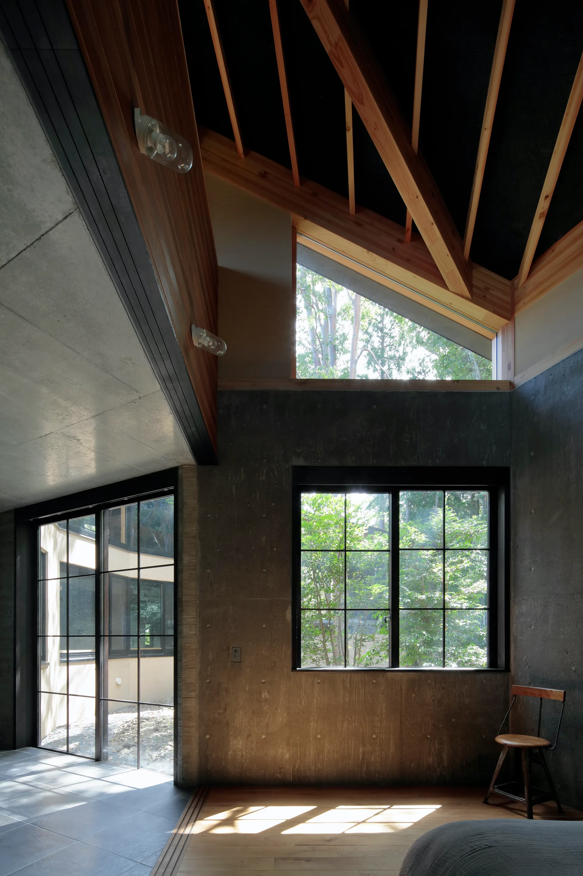 A modern residential interior featuring raw grey concrete walls, large black-framed grid windows, and a high ceiling with exposed wooden rafters.