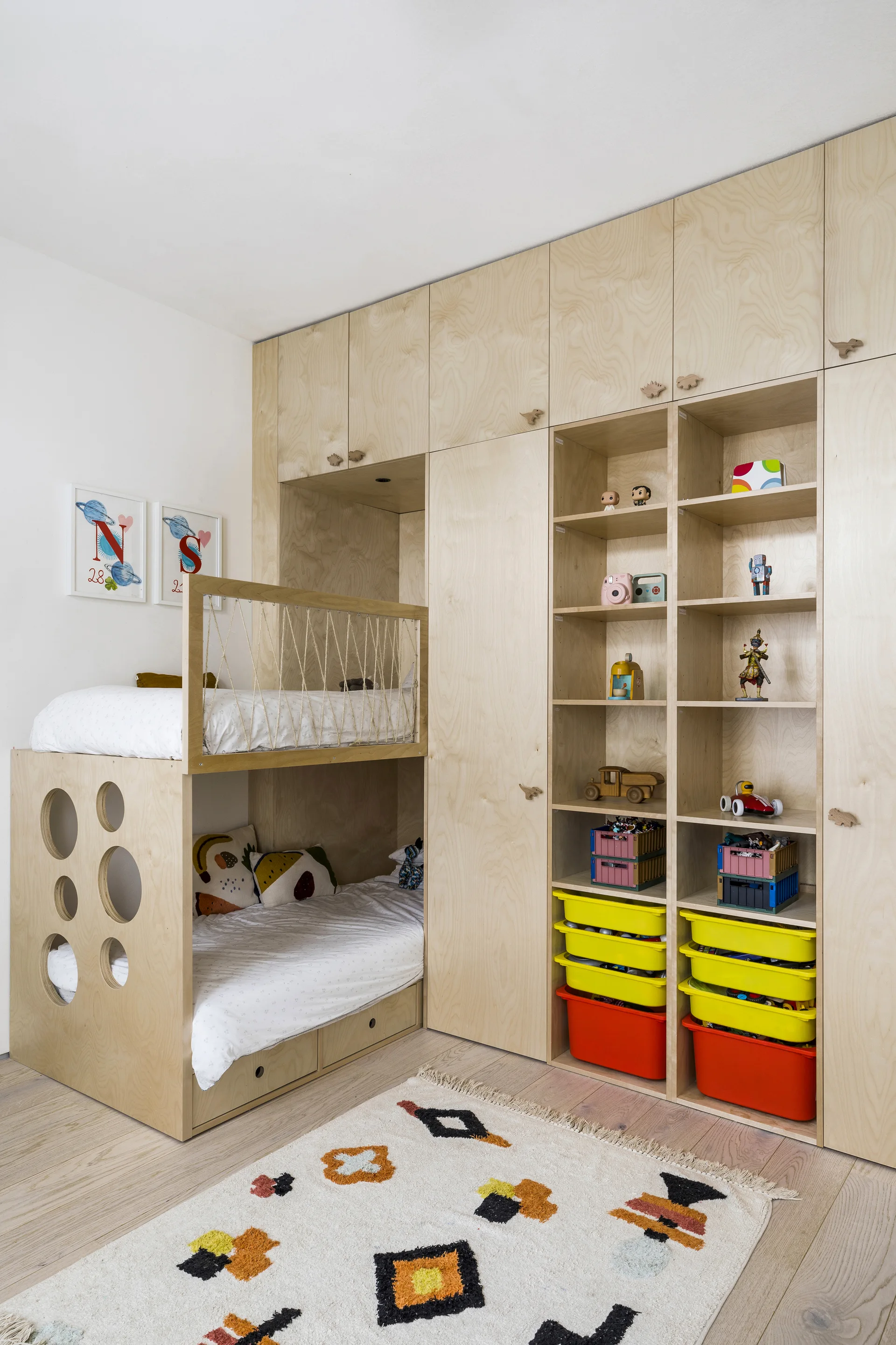 A kids bedroom featuring a custom plywood bunk bed built into a wall of matching cabinets, open toy shelves, and a patterned rug.