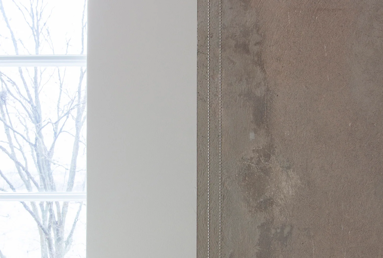 A close-up of a textured grey plaster wall detailed with two vertical lines of silver nailhead trim next to a smooth white painted partition.