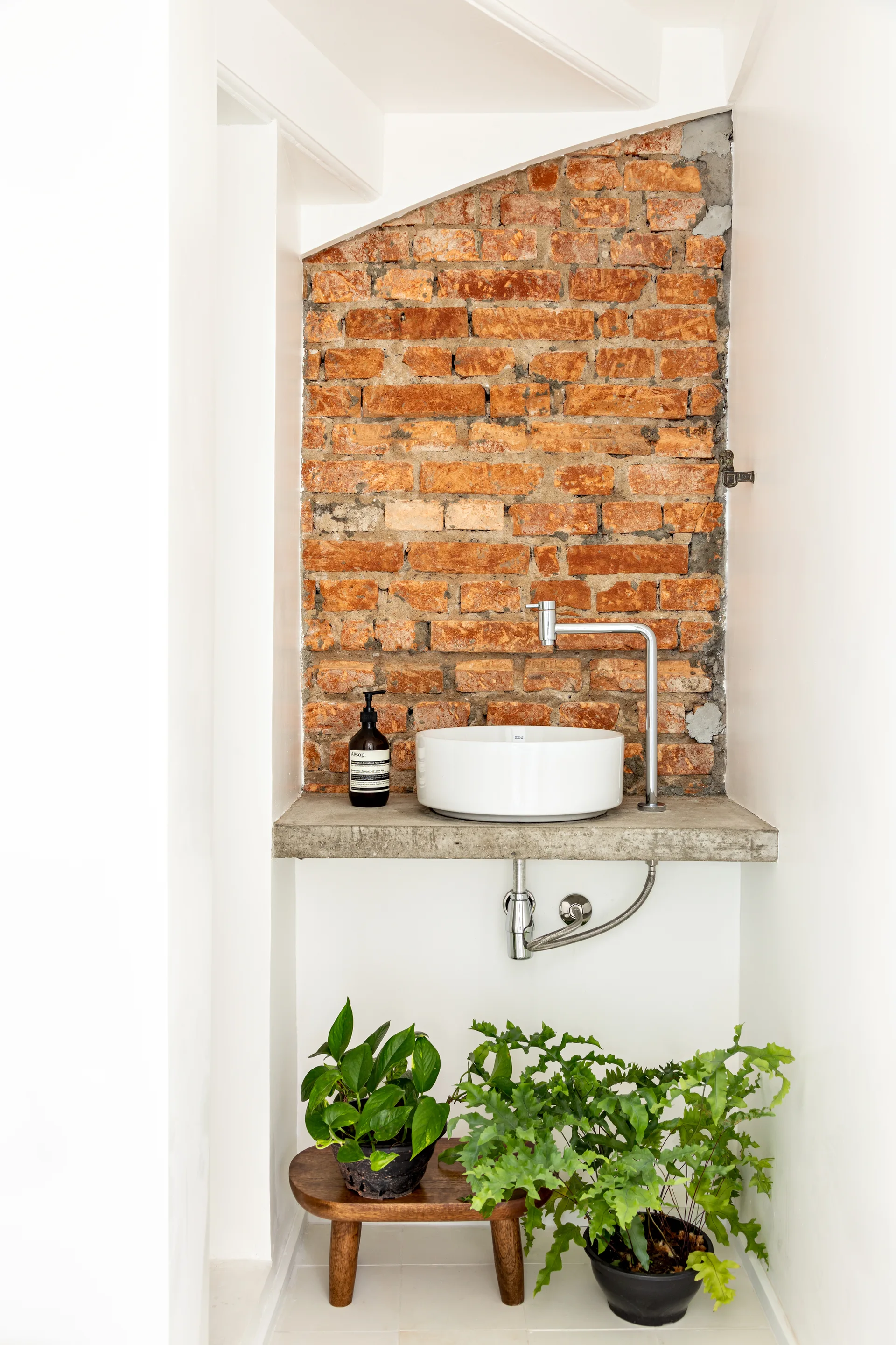 A white vessel sink rests on a concrete shelf beneath a staircase, backed by an exposed red brick wall with potted plants below.