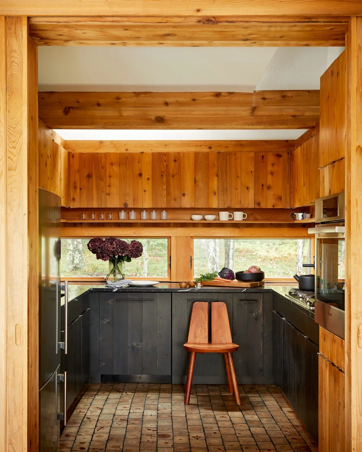 A rustic kitchen featuring dark lower cabinets, warm vertical wood paneling, horizontal windows looking out to trees, and a wooden chair.