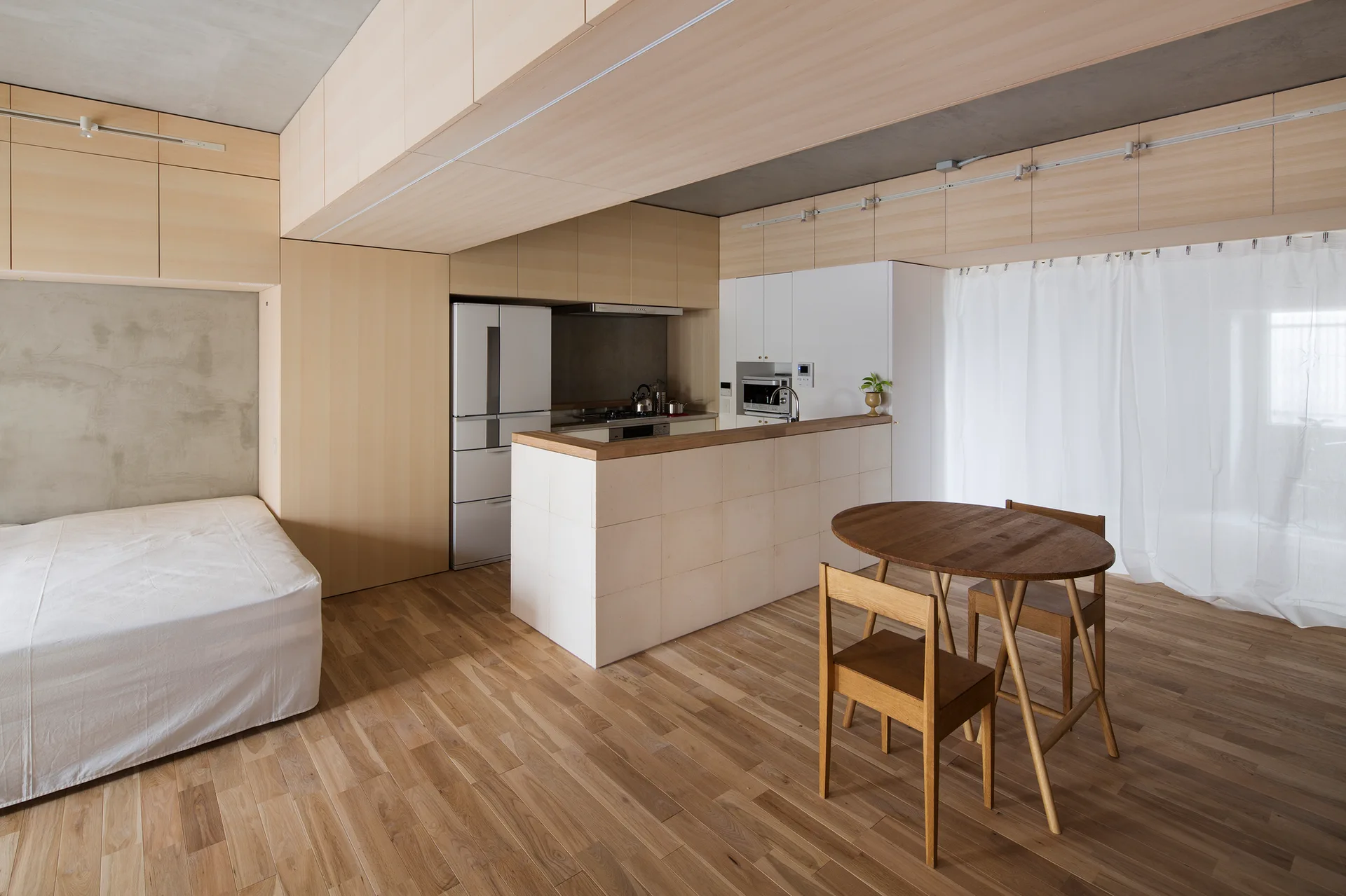 A minimalist studio apartment featuring light wood cabinetry, a tiled kitchen island, a wooden dining set, and an exposed concrete ceiling.