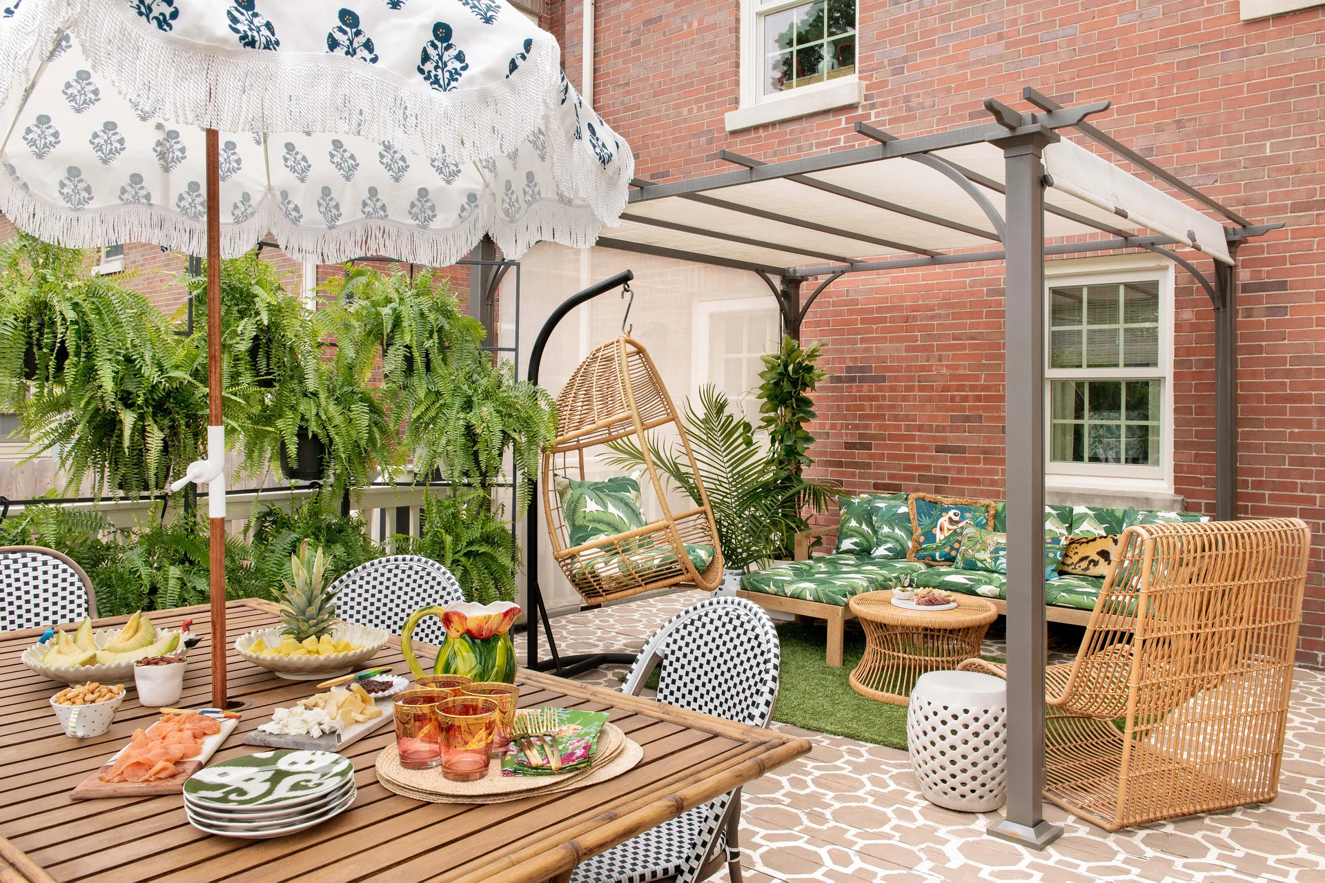 A brick patio features a wooden dining table under a fringed umbrella and a lounge area shaded by a metal pergola with botanical-print cushions.