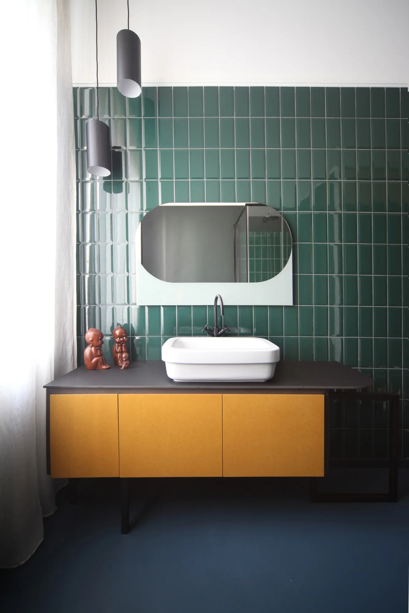 A modern bathroom features a mustard-yellow vanity with a white vessel sink against a glossy dark green tiled wall and an oval wall mirror.