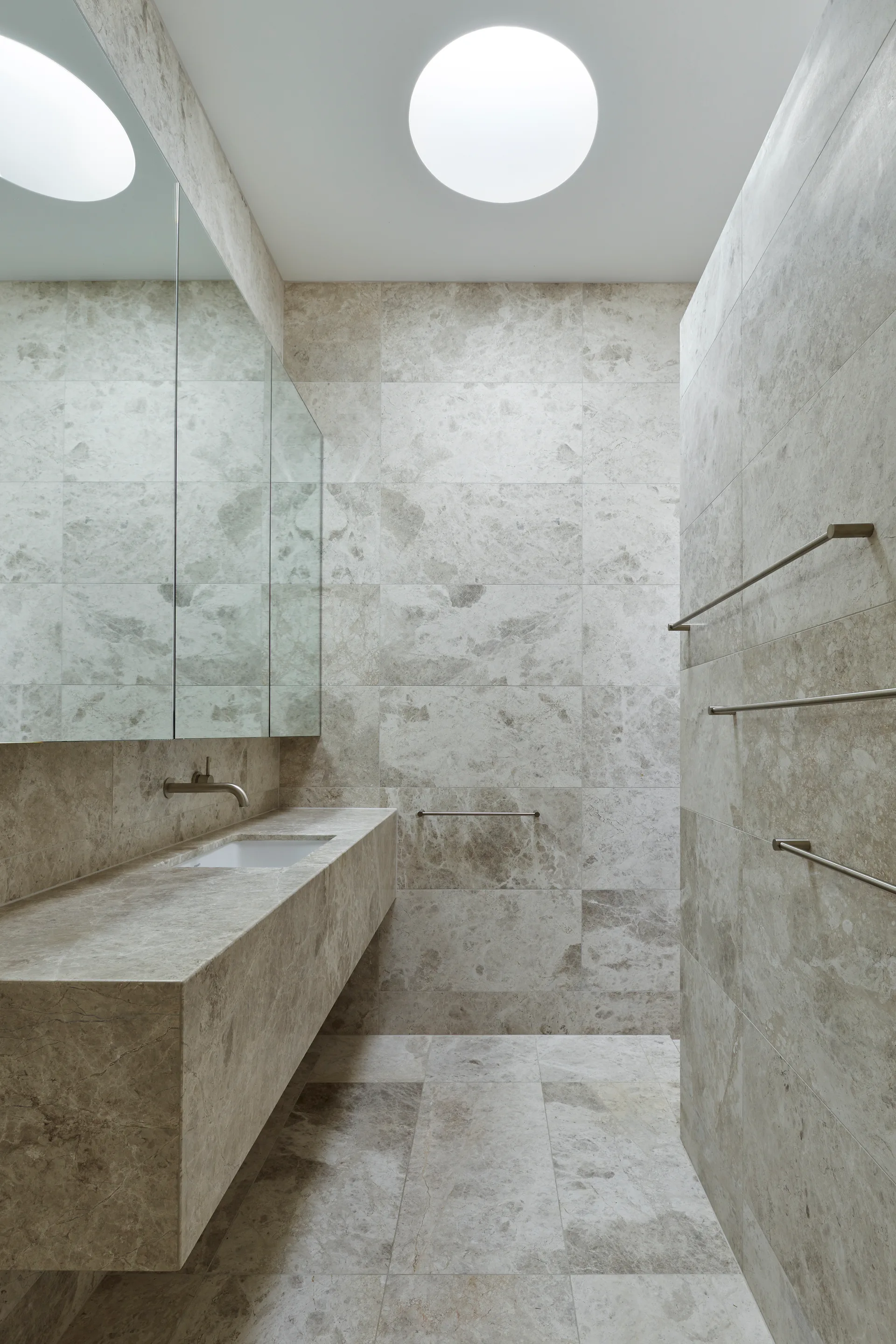 A minimalist bathroom featuring floor-to-ceiling gray stone tiles, a floating stone vanity, and a circular skylight overhead.