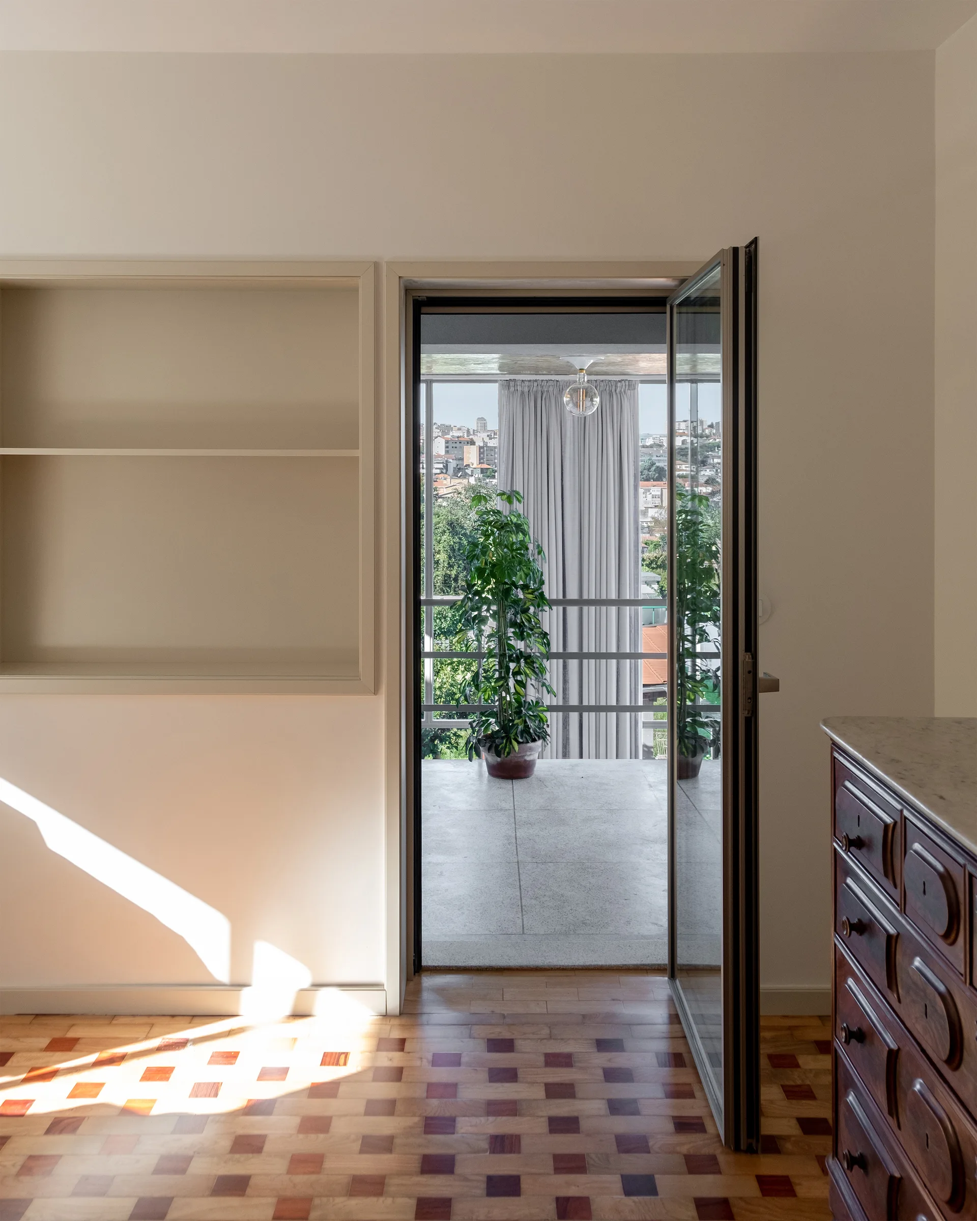 A bright room with geometric wood parquet flooring features a built-in wall shelf and an open glass door leading to a balcony with a potted plant.