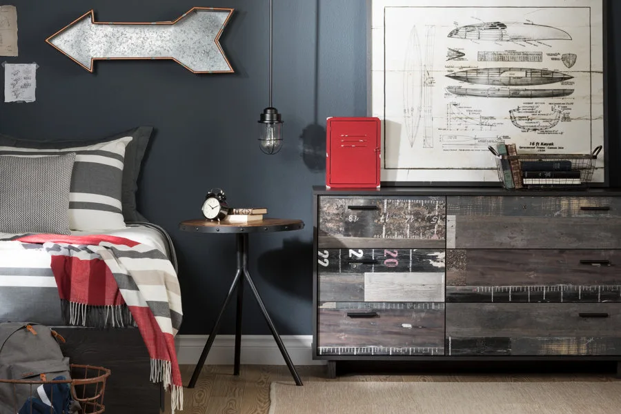 A bedroom corner features a bed with striped bedding next to a rustic wood dresser and a dark grey wall decorated with a metal arrow sign.