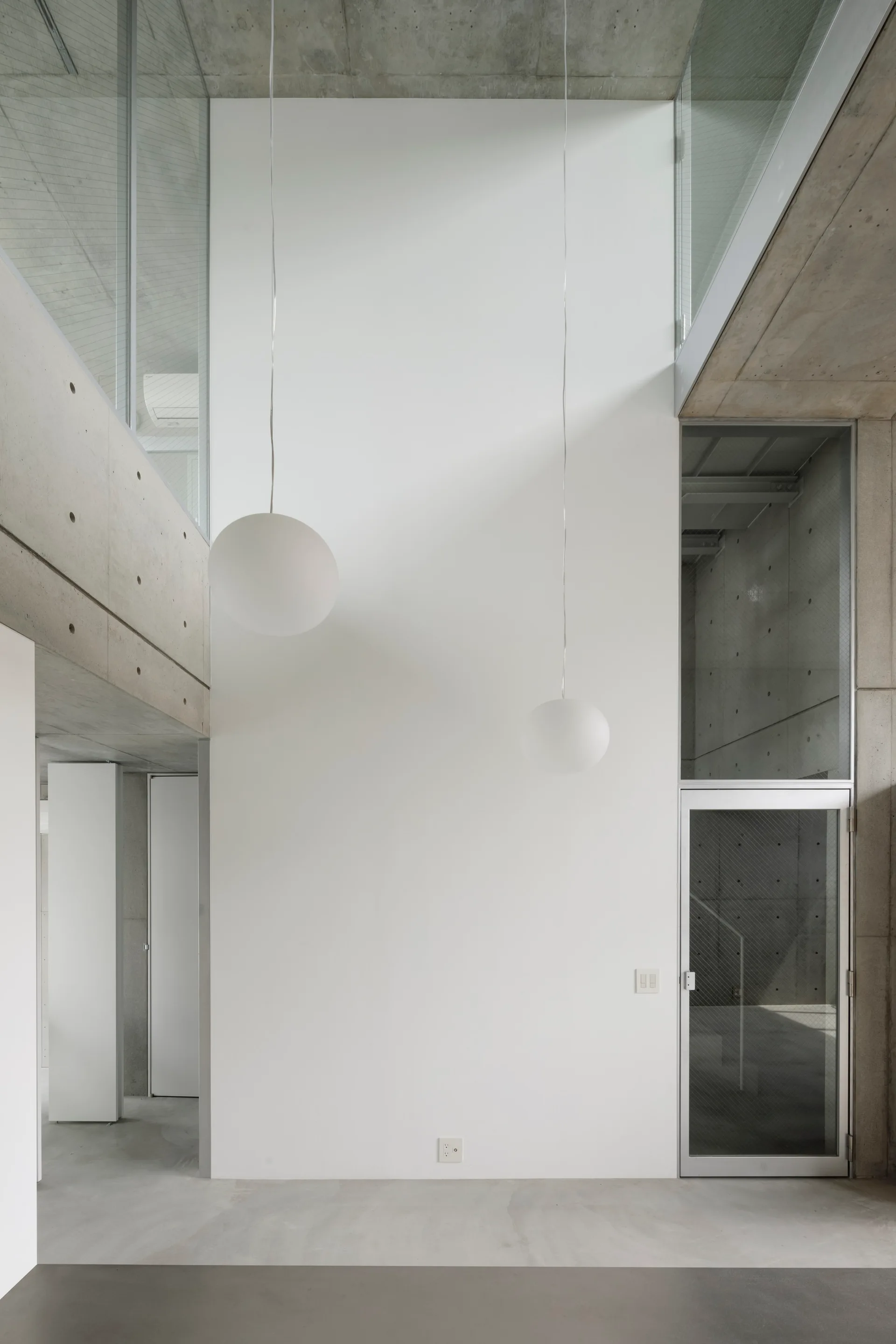 A minimalist interior features a tall white wall flanked by exposed concrete, with two white sphere pendant lights hanging from the ceiling.