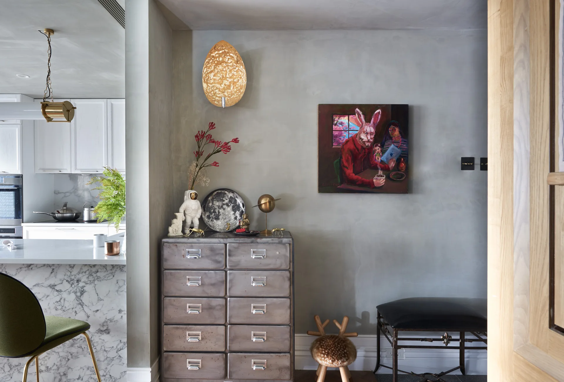 An industrial metal chest of drawers stands against a textured grey plaster wall, styled with whimsical decor under a gold wall sconce.