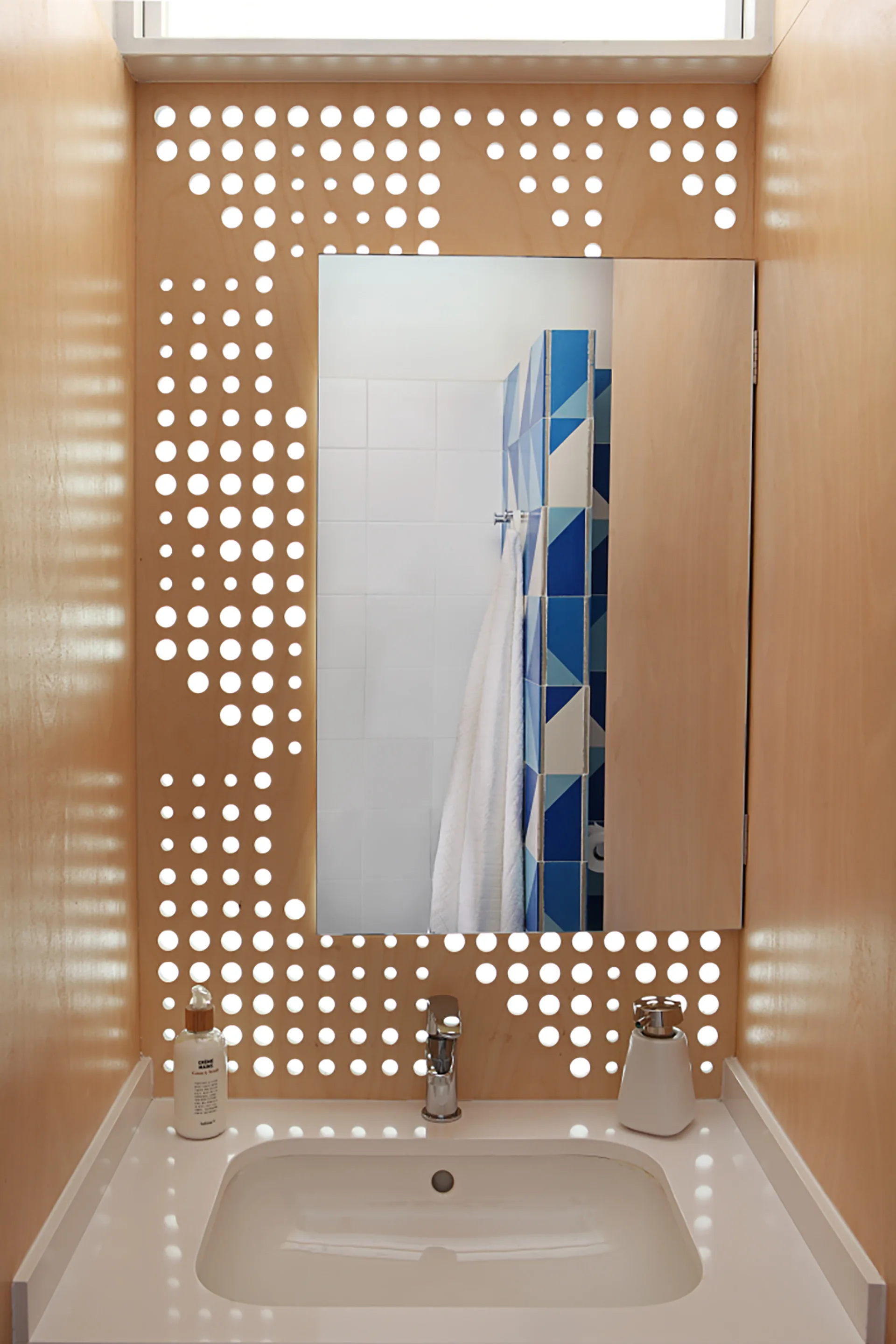 A modern bathroom vanity features a white sink, a chrome faucet, and a rectangular mirror mounted on a perforated light wood plywood panel.