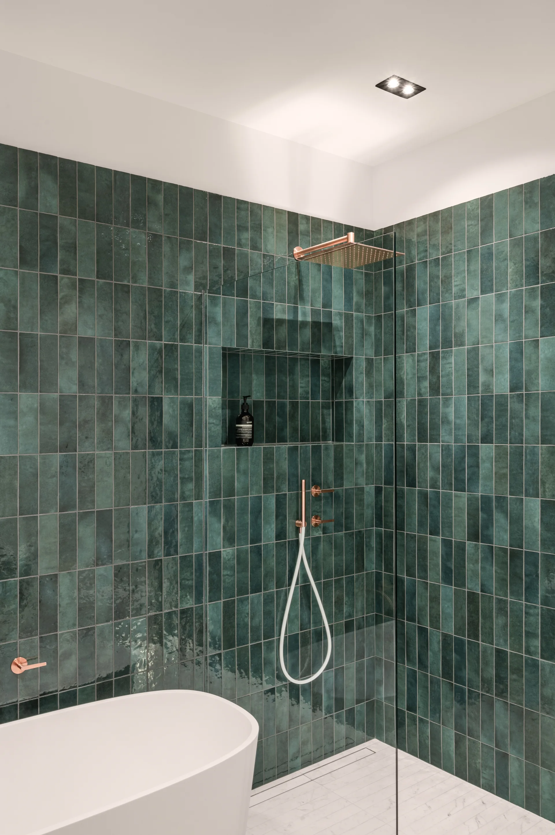A bathroom shower featuring dark green vertical subway tiles, copper fixtures, a clear glass partition, and a white freestanding bathtub.
