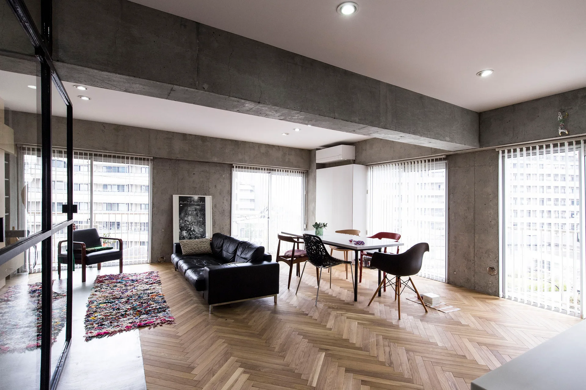 An open-concept living room features exposed concrete walls and ceiling beams, a black leather sofa, and a warm herringbone wood floor.