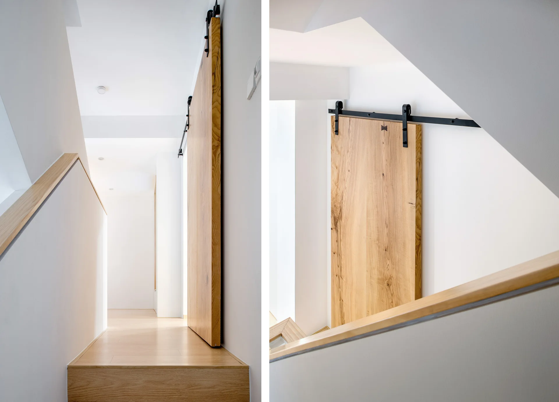 A light wood sliding barn door on a black metal track is mounted on a white wall above a staircase landing with a wooden handrail.