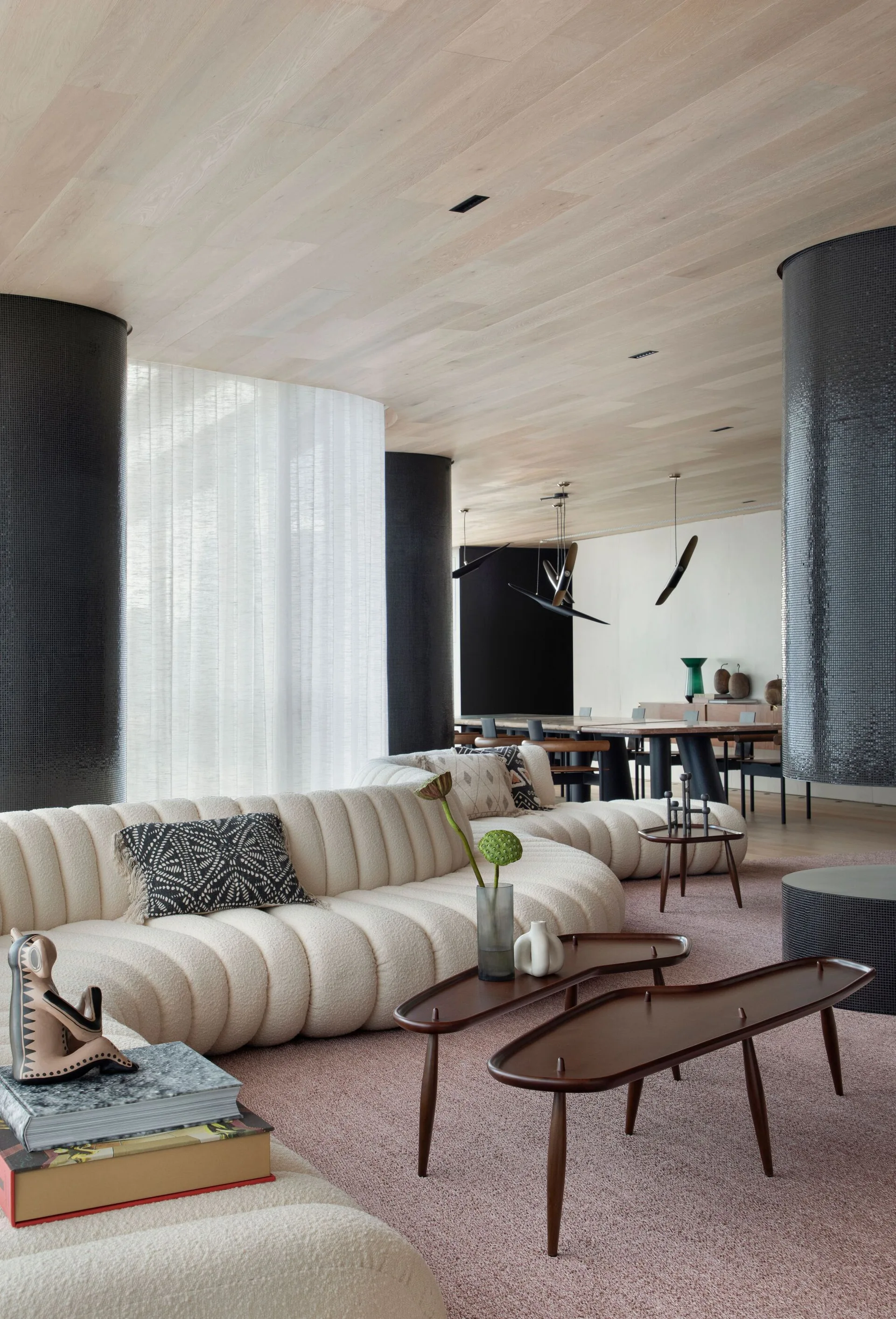 A contemporary living room featuring a curved cream-colored sofa, dark tiled columns, and organic wood coffee tables set under a light wood ceiling.