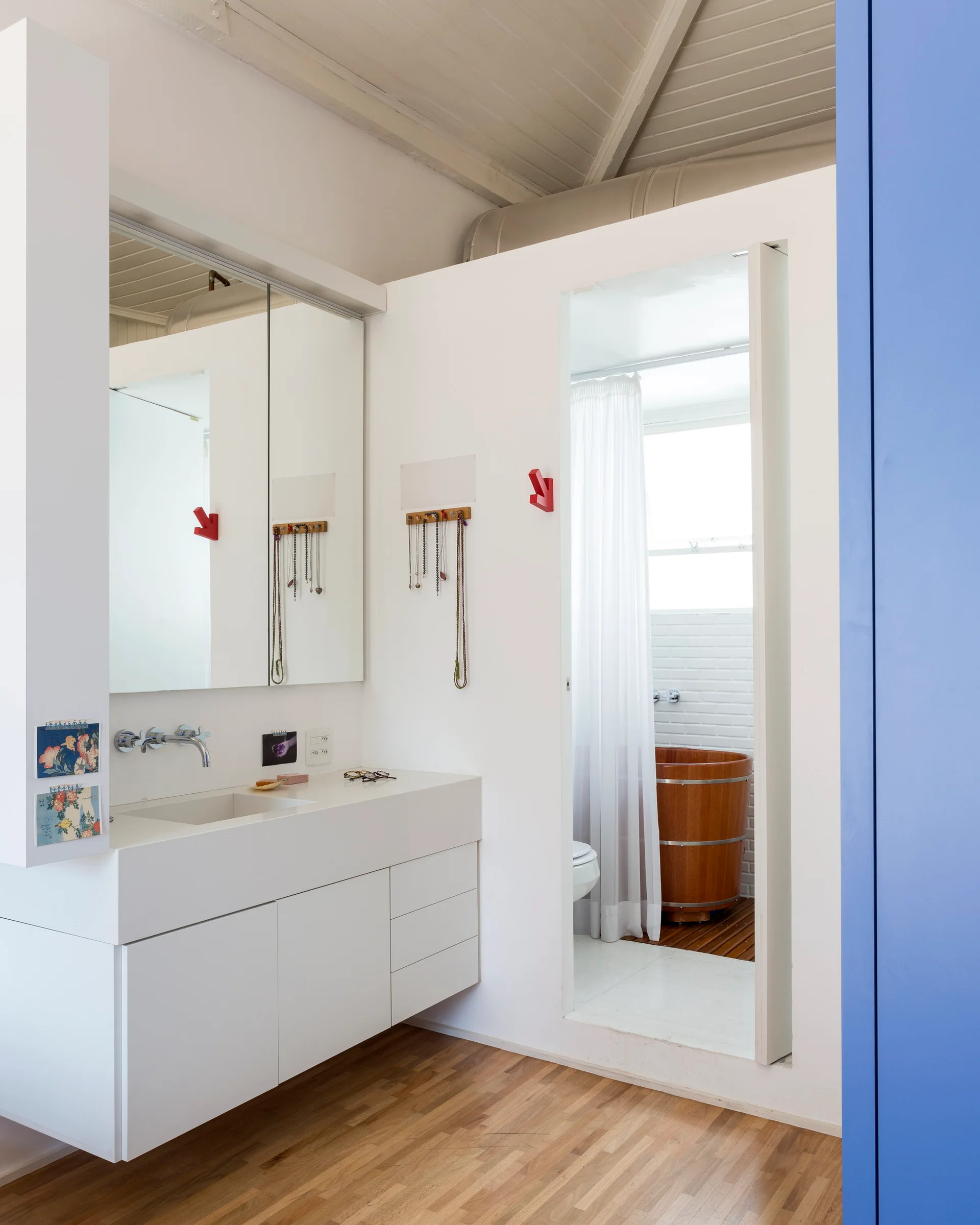A minimalist bathroom featuring a white wall-mounted vanity, a large mirror, and an open doorway revealing a cylindrical wooden soaking tub.