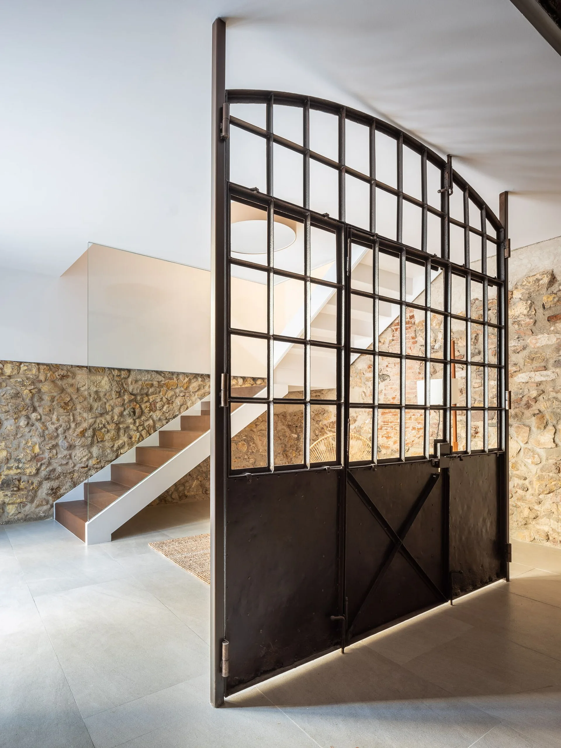 An industrial black metal partition stands on a grey tile floor near a modern wooden staircase with a glass railing and rustic stone wall.