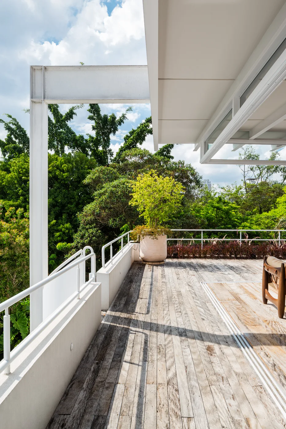 An elevated outdoor wooden deck features a white metal railing system, structural overhead beams, and a large planter against lush green trees.