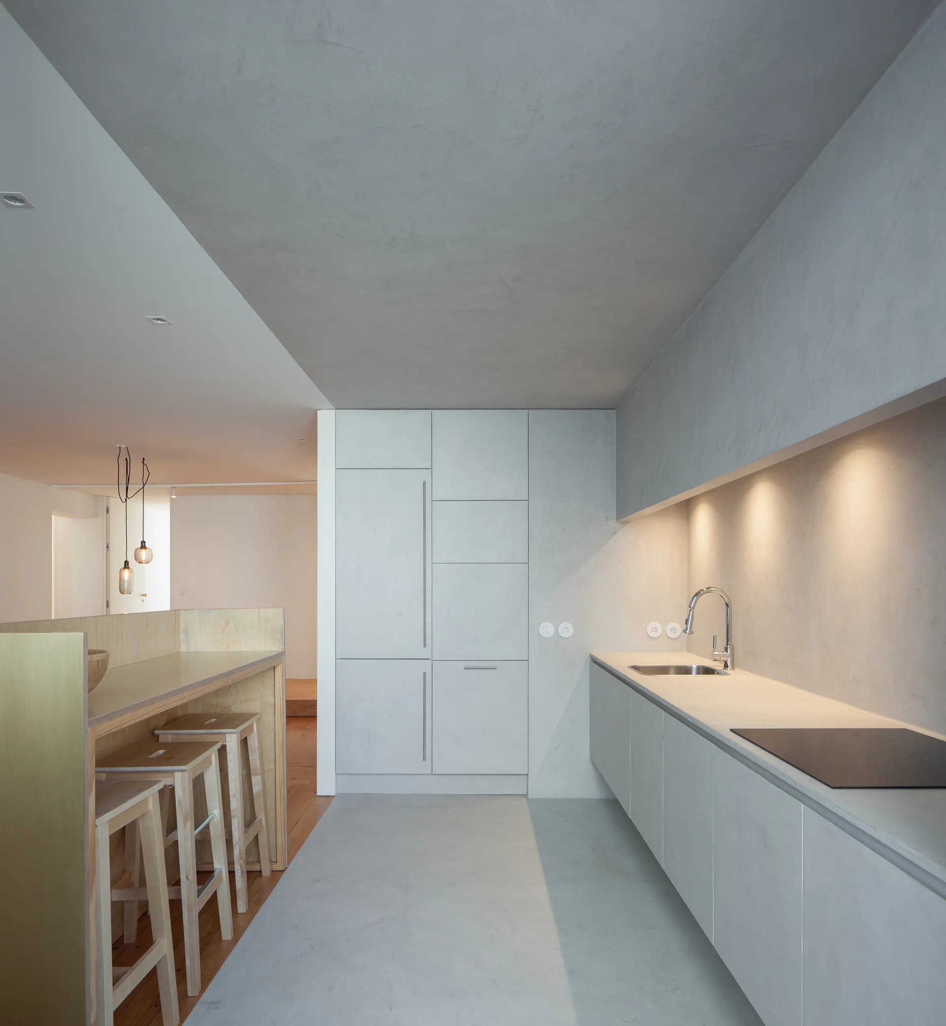 A minimalist kitchen featuring seamless gray plaster cabinets, a stainless steel sink, and a light wood breakfast bar with matching stools.