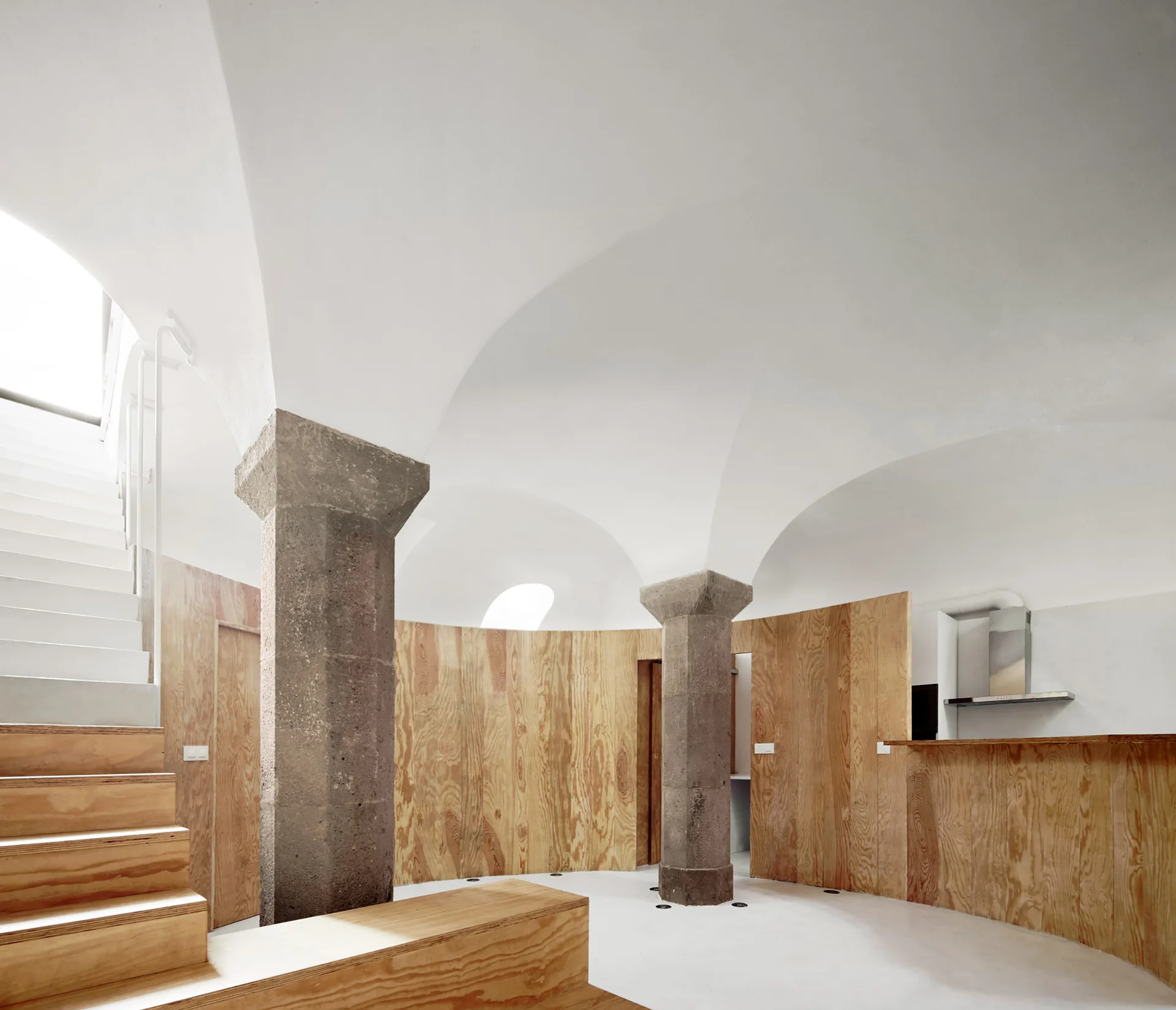 A white vaulted ceiling interior featuring rustic stone pillars, curved plywood wall paneling, and a wooden staircase.