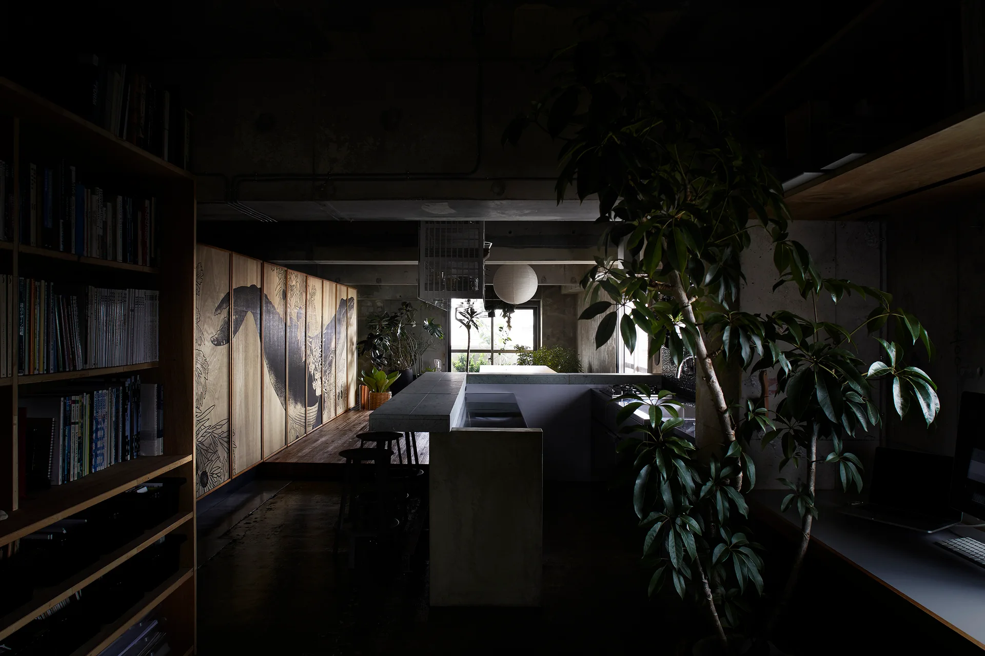 A dimly lit industrial kitchen features a concrete island, tall wooden bookshelves, and large wooden cabinet doors adorned with a black wave mural.